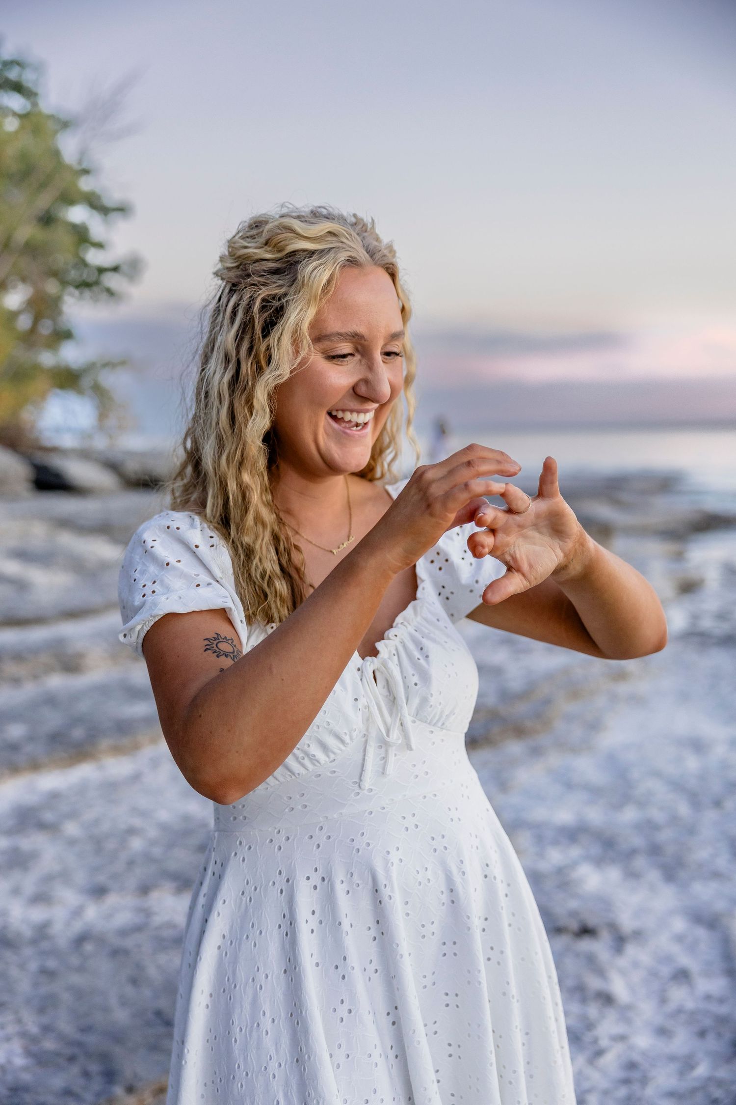 Happy blonde woman in white dress admiring engagement ring on rocky beach at sunset.