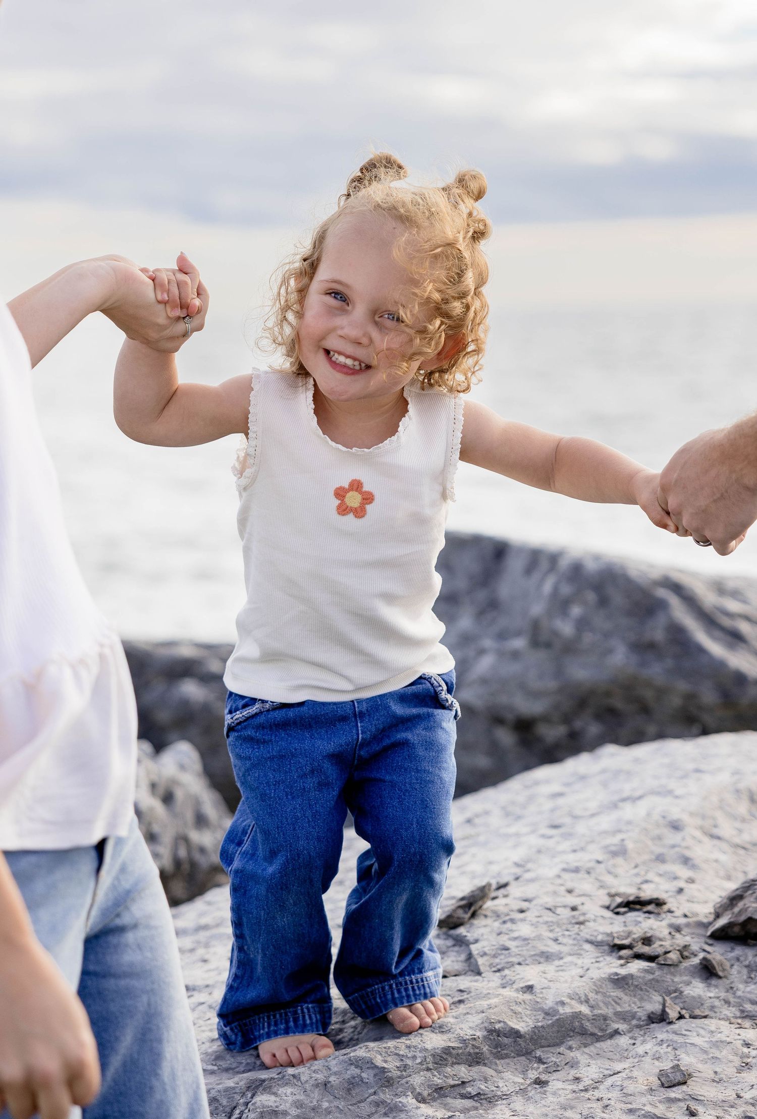 Smiling toddler girl with curly hair in white tank top and jeans standing on rocks at beach holding adults hands.