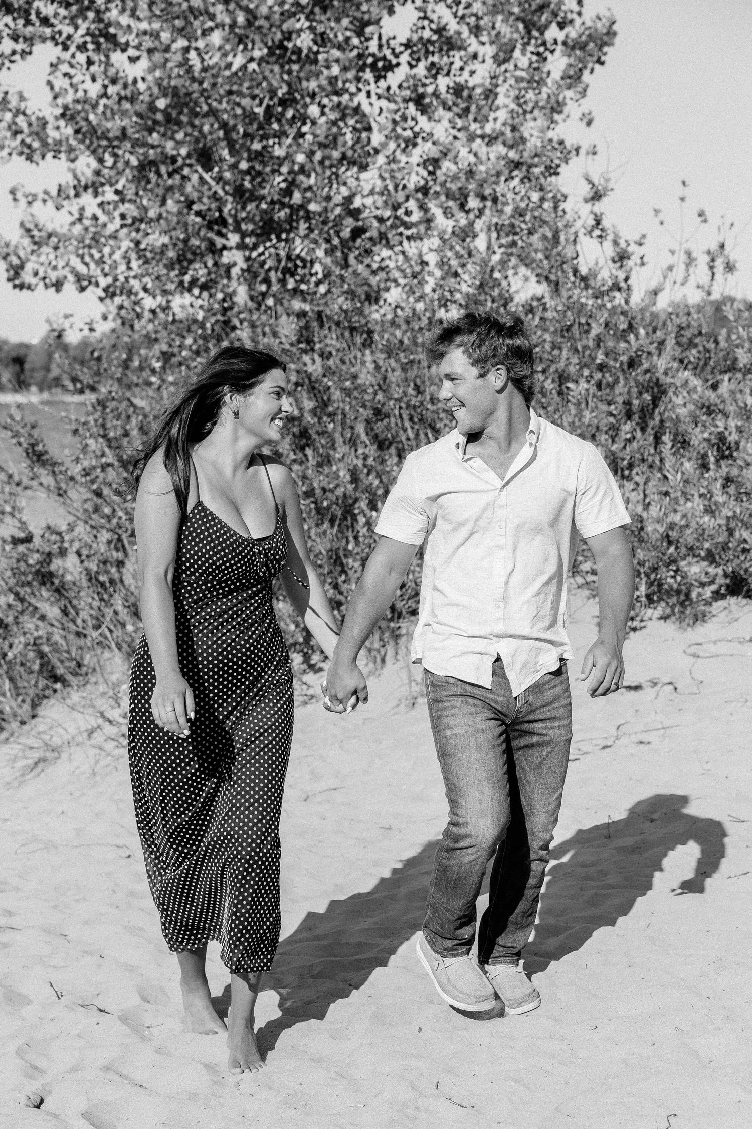 Black and white photo of a couple holding hands and smiling at each other while walking on a sandy beach path.