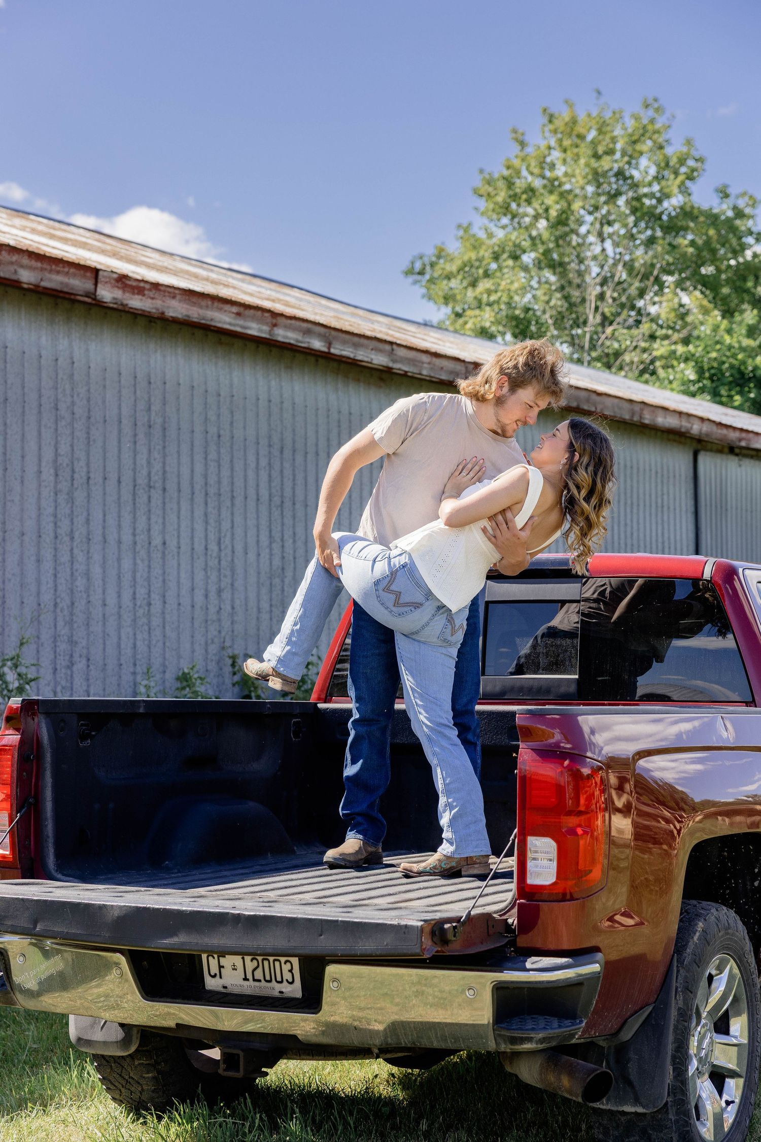 Couple sharing a romantic dip kiss while standing in the bed of a red pickup truck beside a rustic barn.