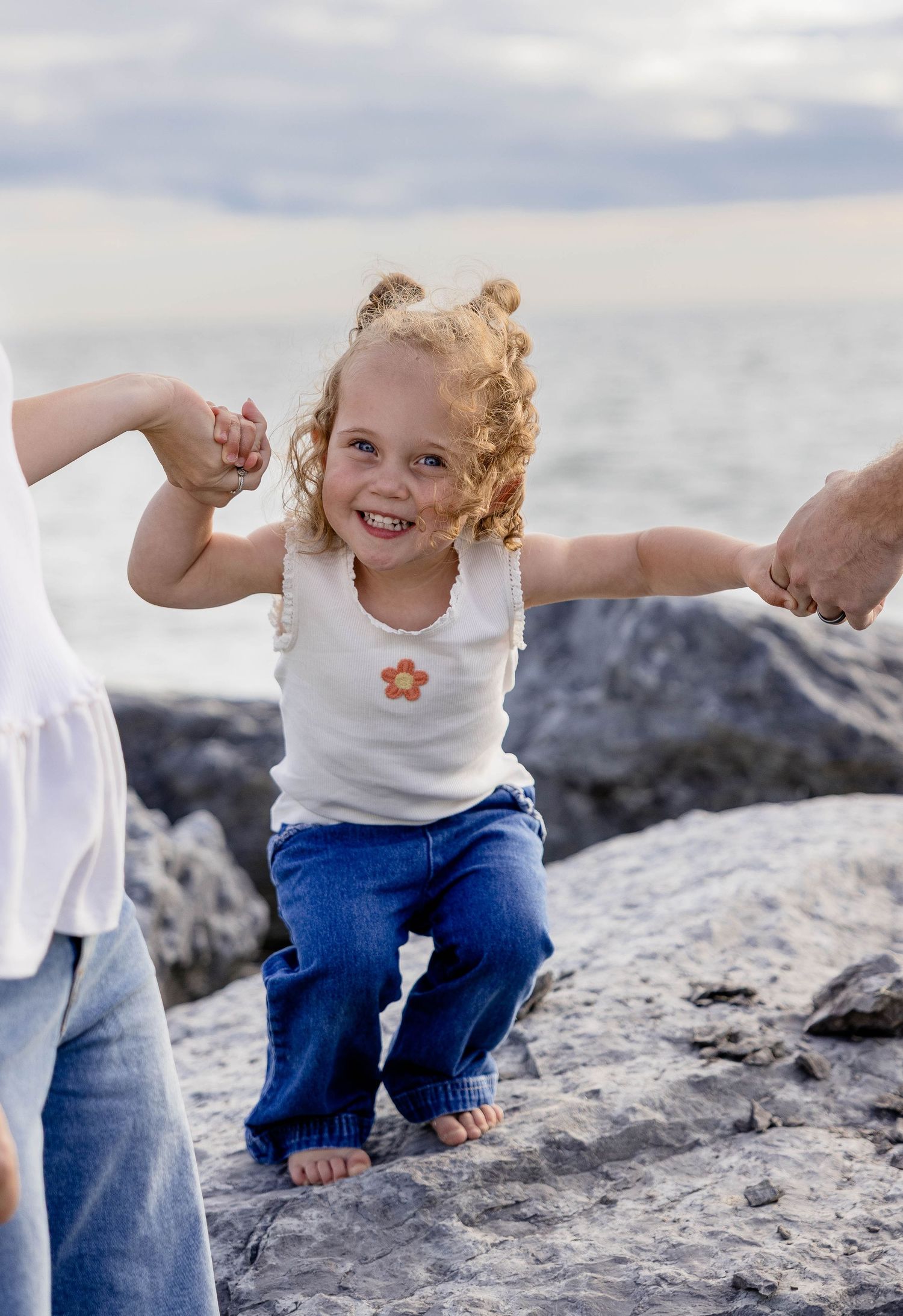 Smiling toddler girl with curly hair holds adults' hands on rocky beach shore wearing white tank top and jeans.