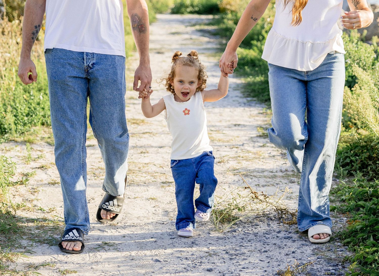 Young toddler girl walks on dirt path holding hands with parents in matching jeans on a sunny day.