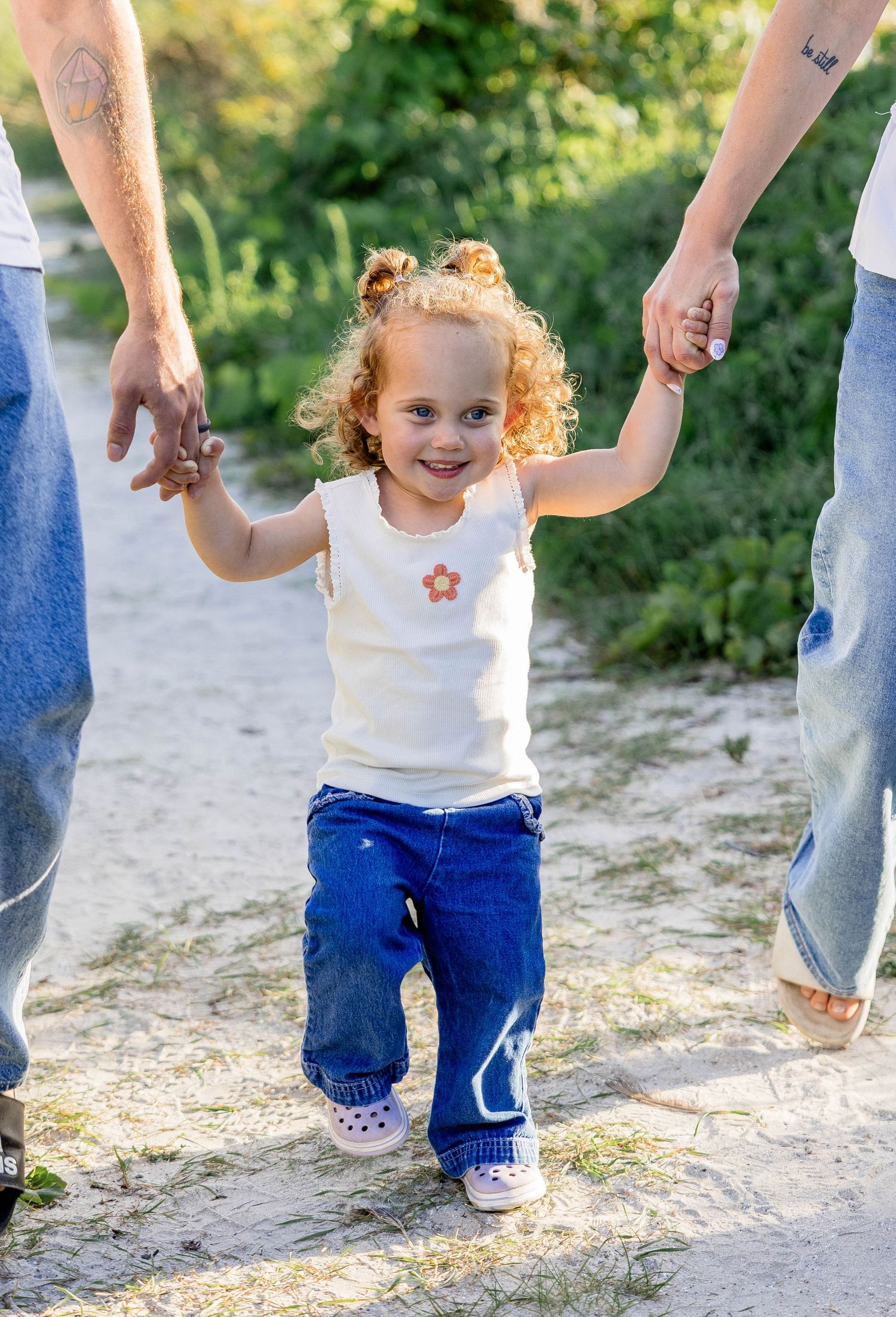 Curly-haired toddler girl smiling while walking outdoors, holding hands with two adults on a sunny day.