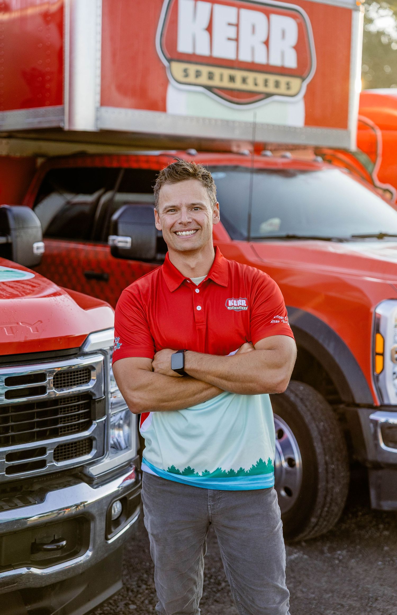 A smiling man in a red Kerr Sprinklers polo stands confidently with arms crossed in front of red company trucks.