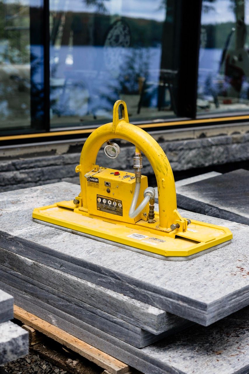 Yellow MQUIP vacuum stone lifter with pressure gauge resting on stacked granite slabs near a glass building.
