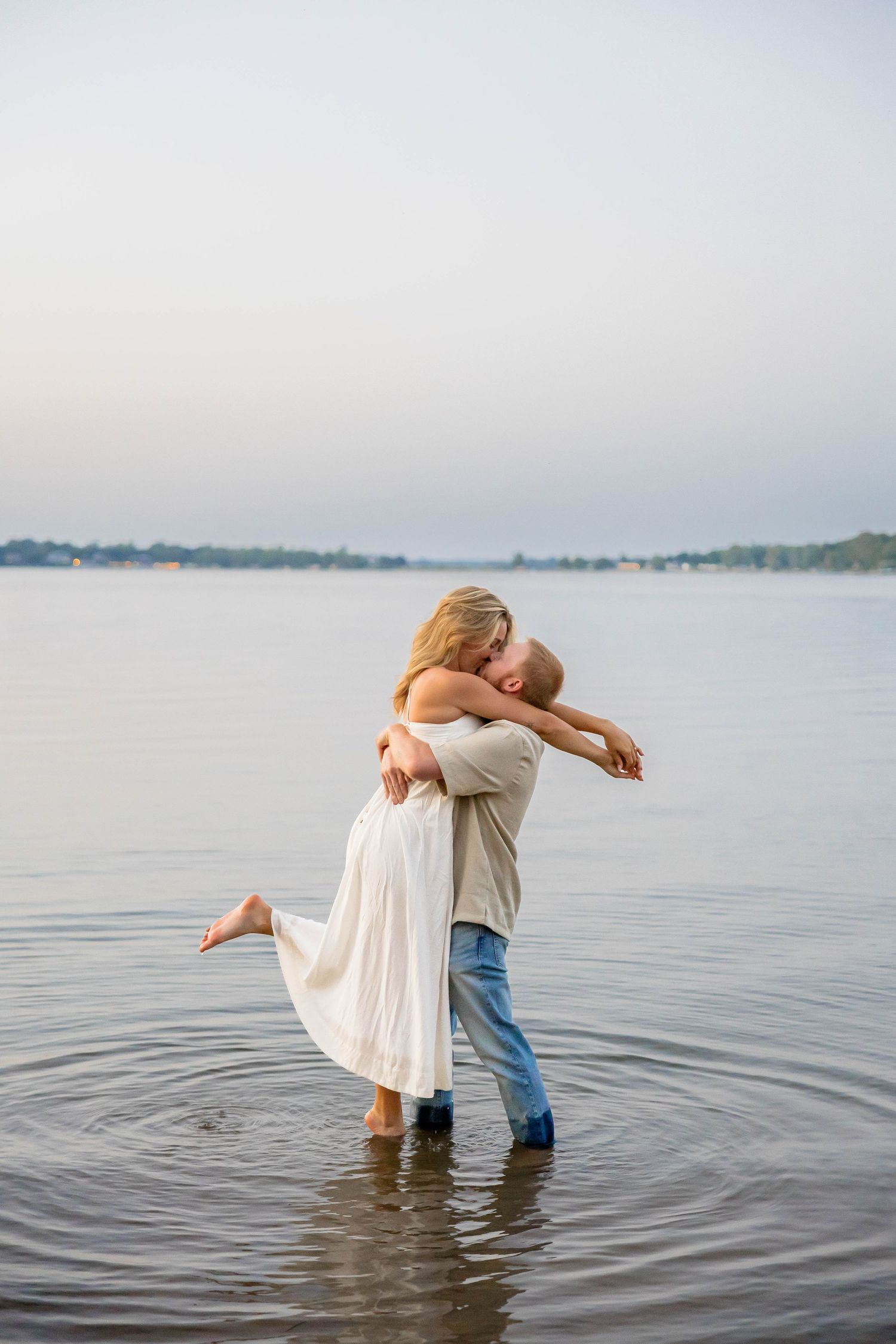 Romantic couple embracing and kissing while standing in shallow lake water, woman in white dress lifted by man in jeans.
