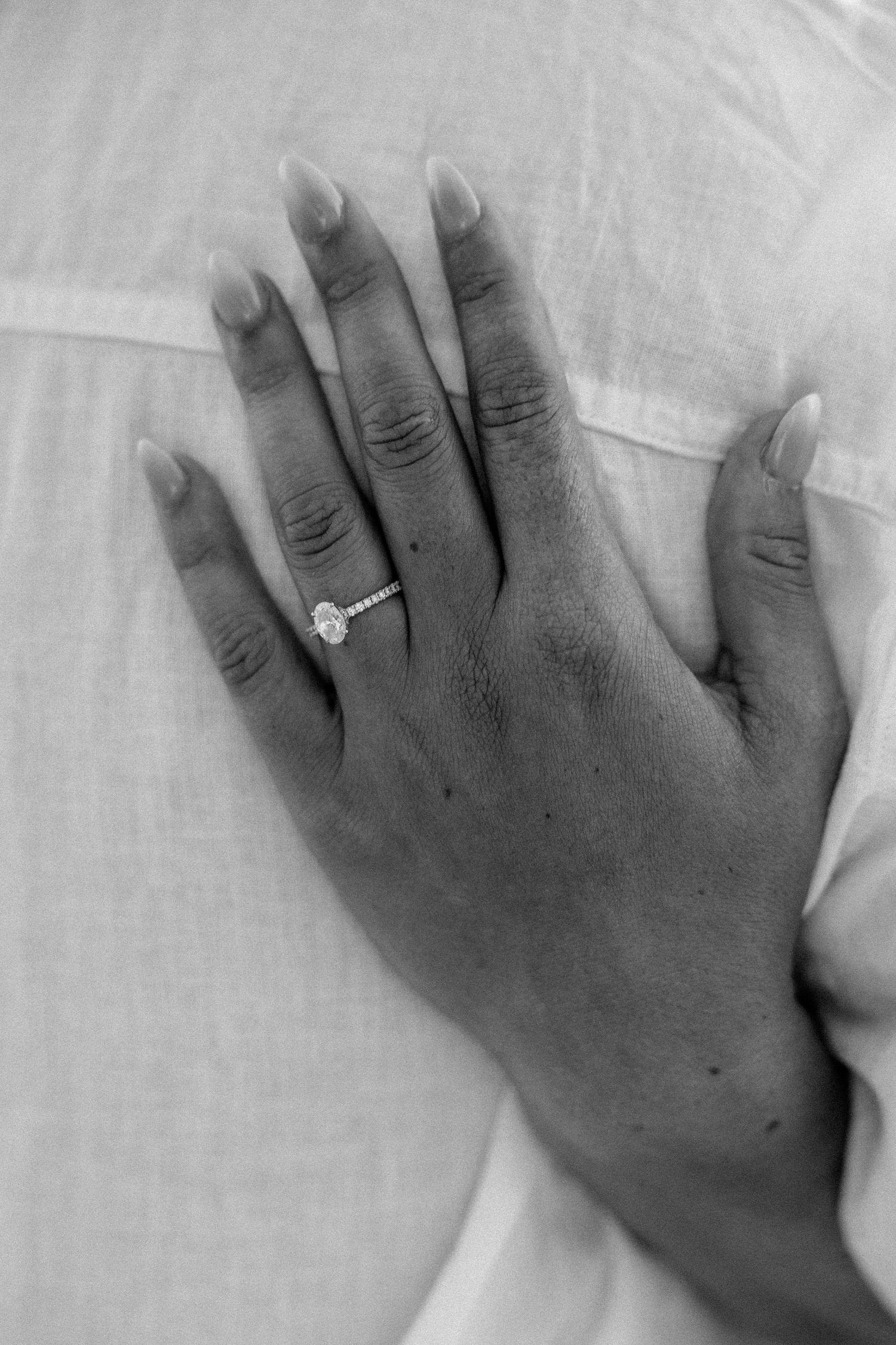 Black and white photo of a woman's hand with an elegant diamond engagement ring resting on a white shirt.