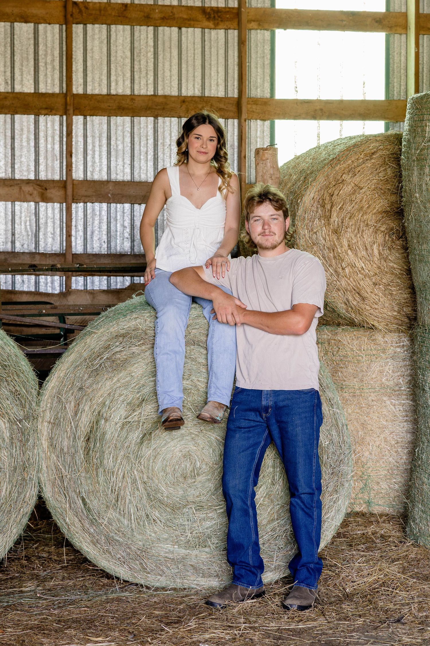 Young couple posing in a rustic barn with large round hay bales, woman sitting on bale, man leaning beside her.