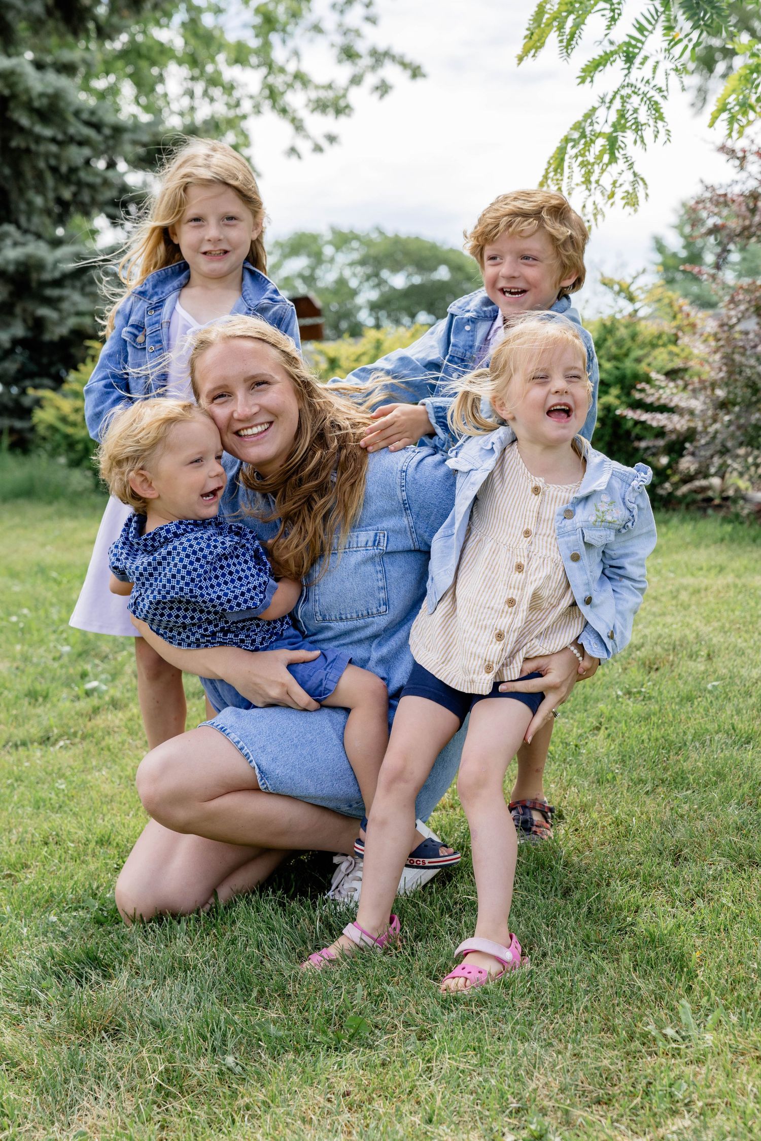 A smiling mother crouches on green grass with four young children in denim outfits, posing together outdoors.