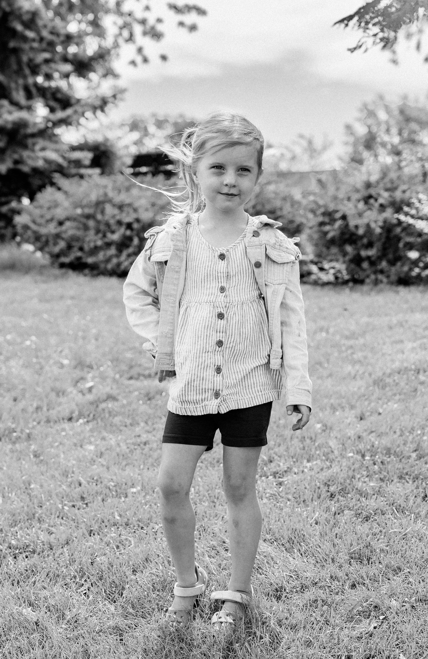Black and white photo of a young blonde girl standing on grass, wearing a button-up shirt and shorts.