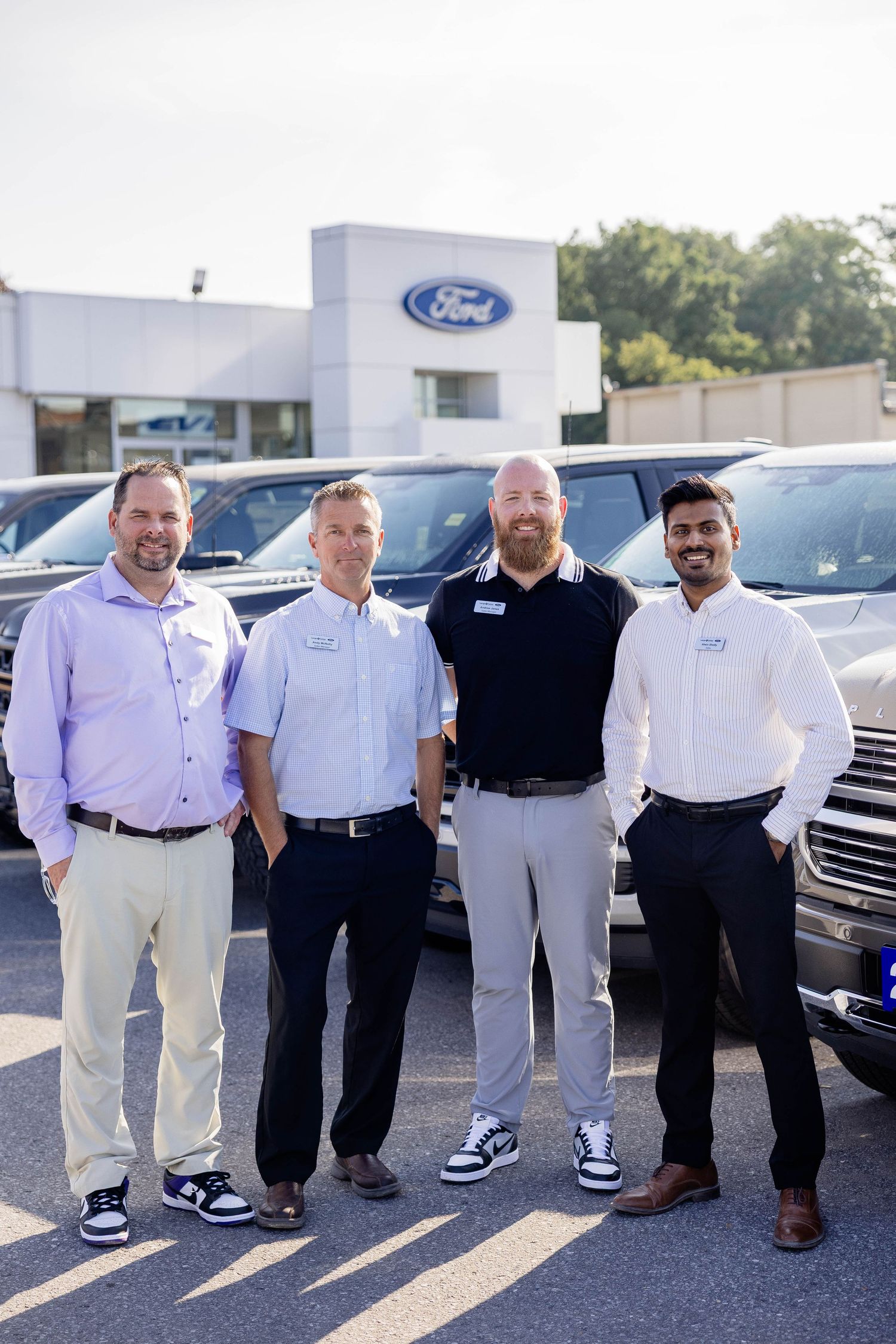 Four Ford dealership employees posing together in a car lot with Ford logo sign visible in background.