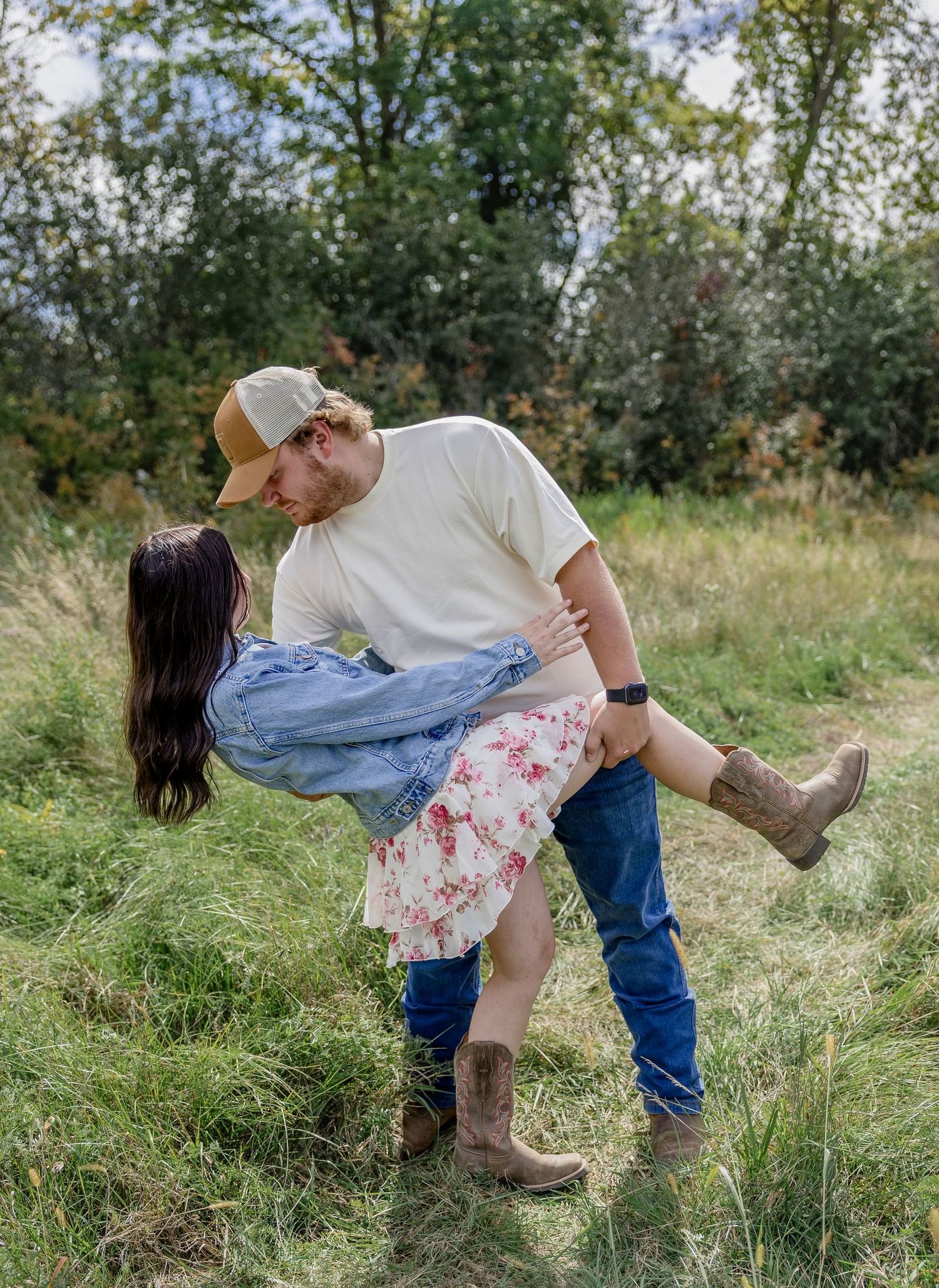 Couple sharing a romantic dip kiss in a grassy field, woman in floral skirt and cowboy boots, man in jeans and cap.