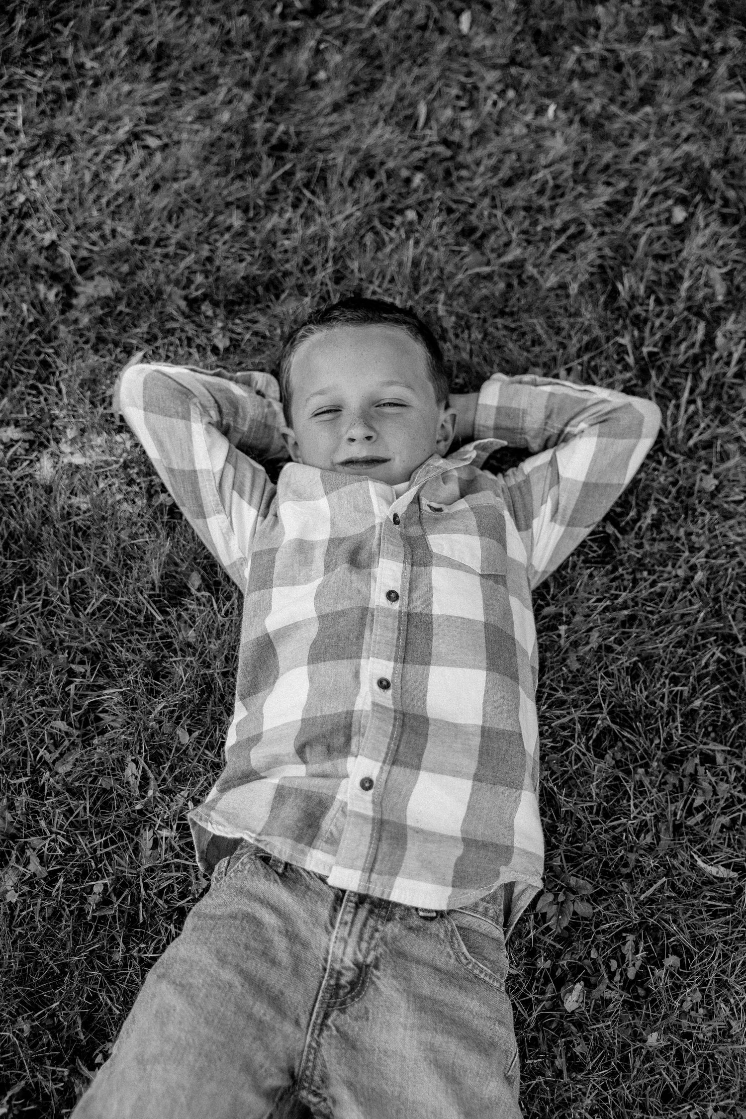 Black and white photo of a young boy lying on grass with hands behind his head, wearing a plaid shirt and jeans.