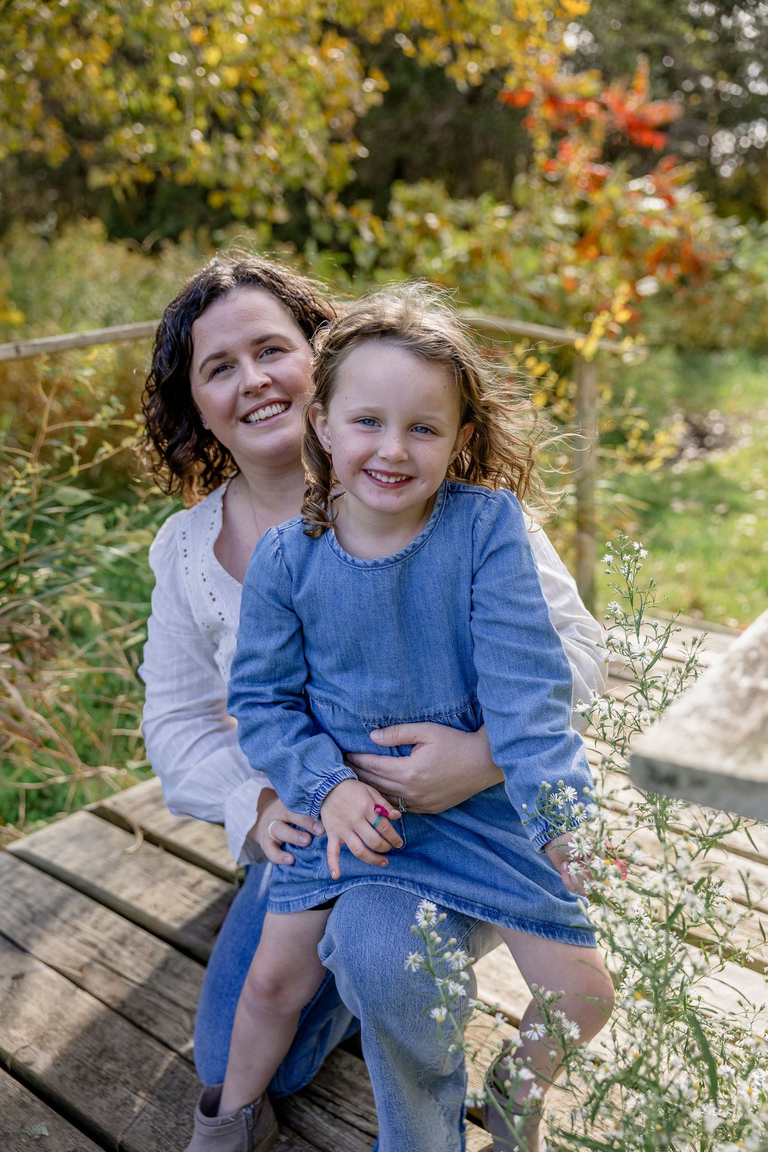 A smiling woman in white holds a young girl in a blue denim dress on a wooden bridge surrounded by autumn foliage.