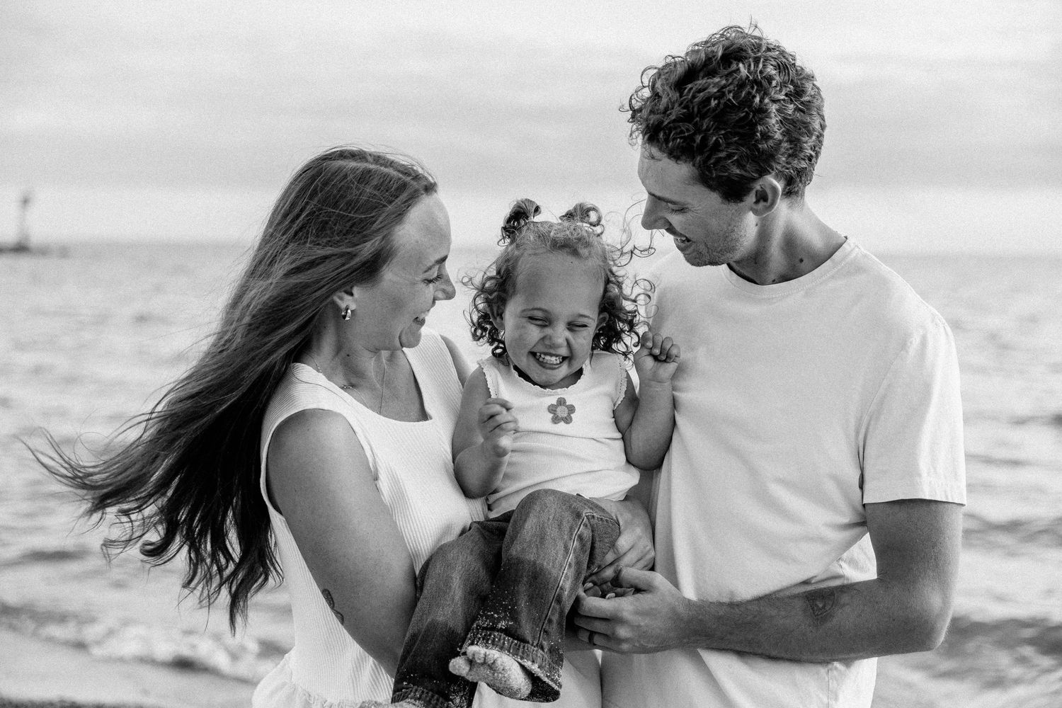 Black and white photo of a happy family with parents holding their laughing toddler girl on a beach.