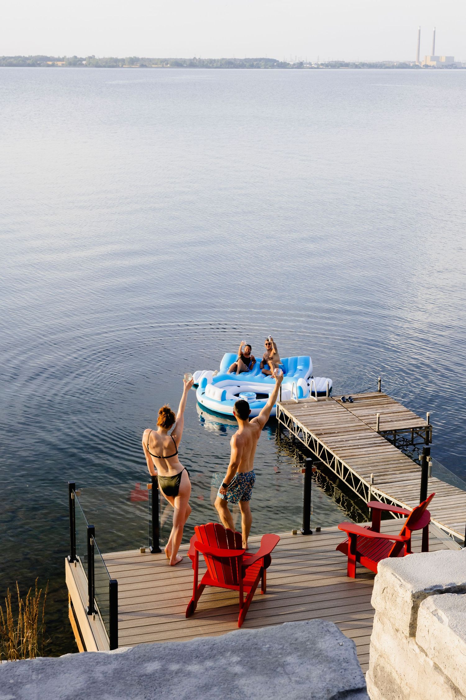 Friends on a wooden dock waving to people on a blue paddle boat on a calm lake.