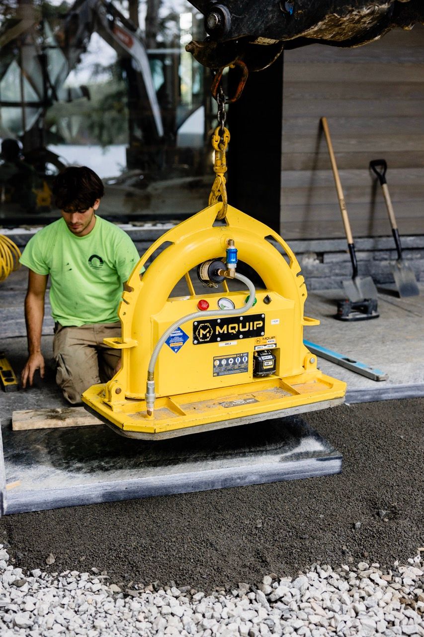 Worker guides yellow MQUIP vacuum lifting device onto concrete slab at construction site.