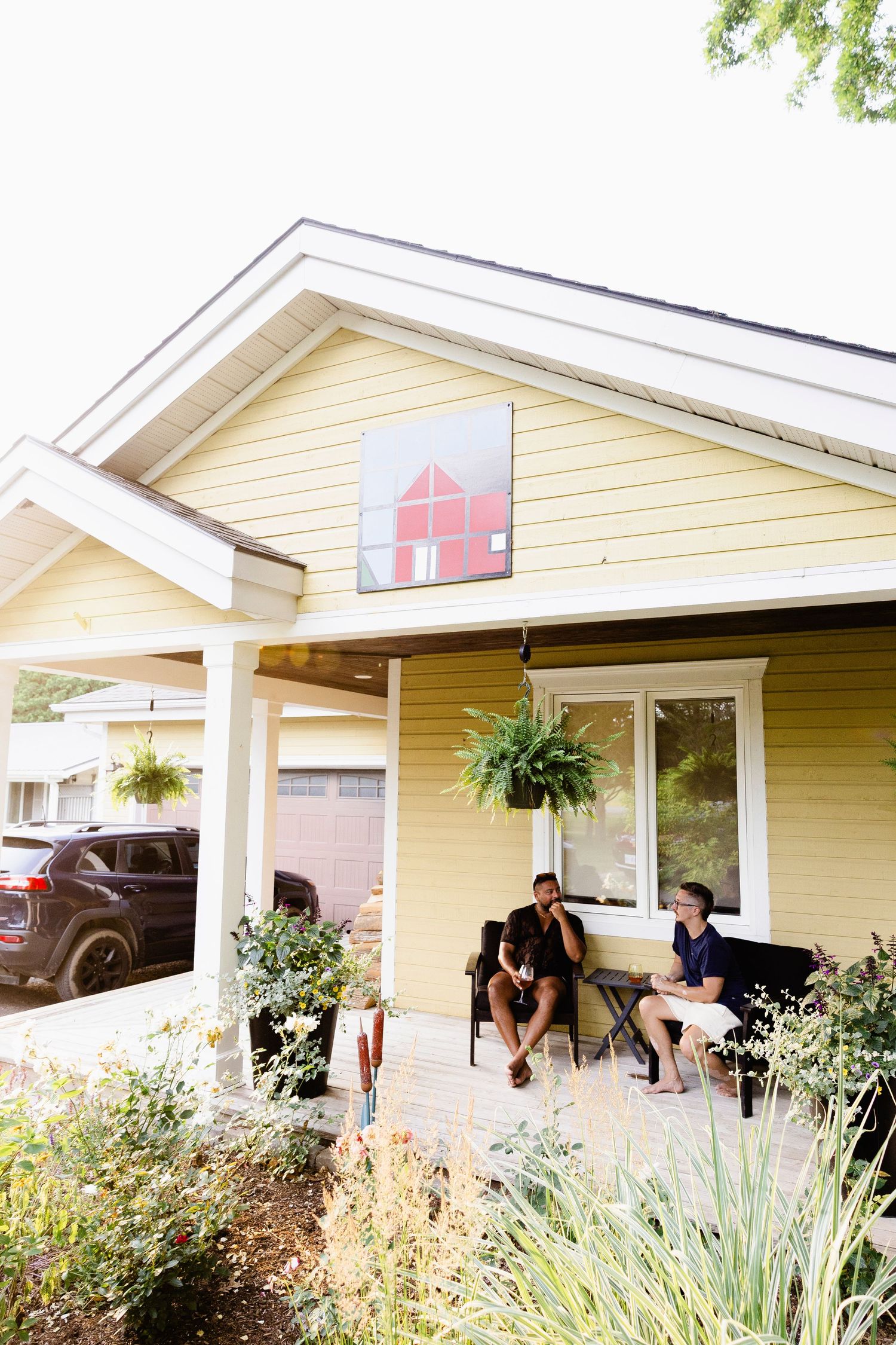 Two people relaxing on porch of yellow house with hanging plants outside.