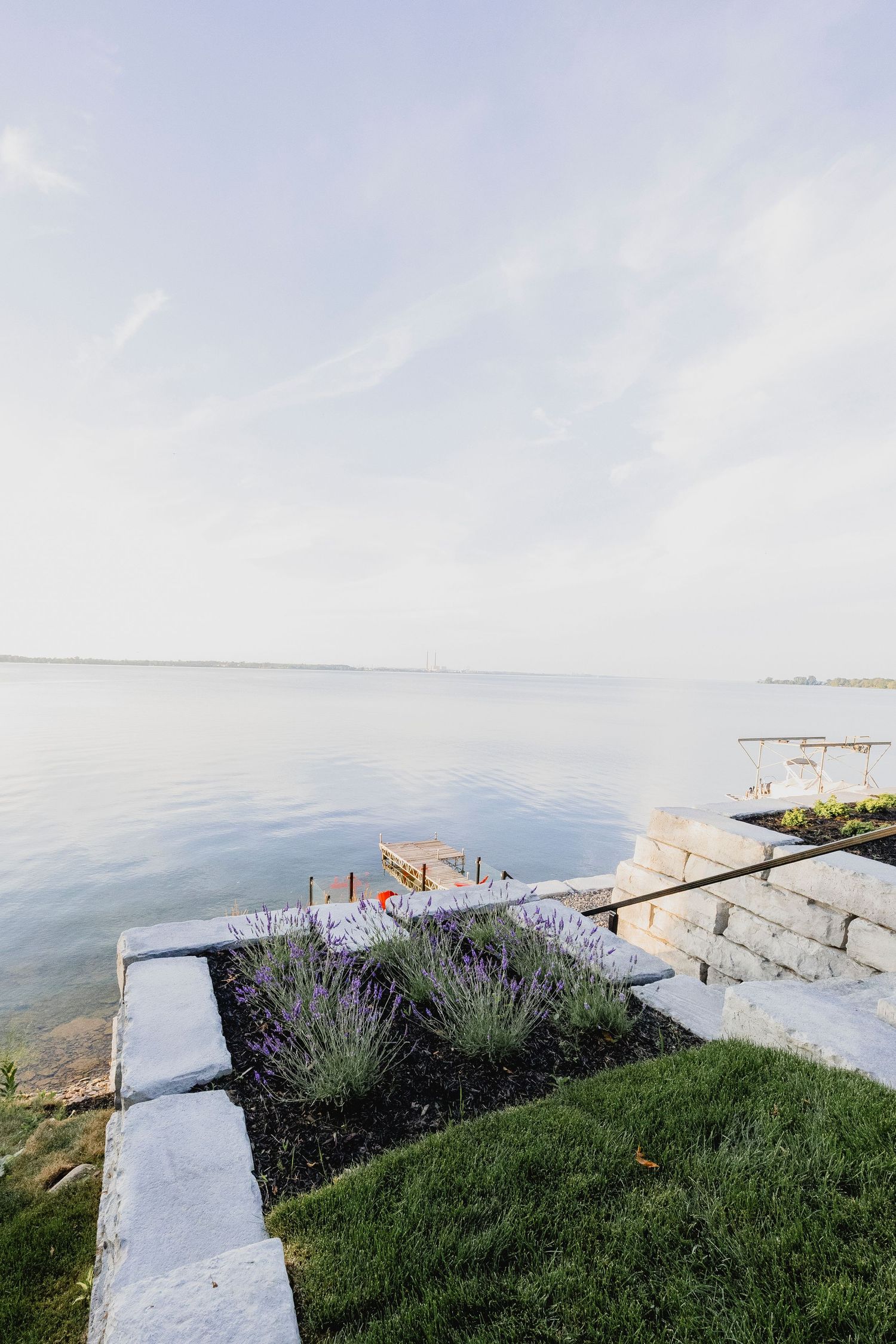 Lakeside stone retaining wall with lavender plants, black mulch, and a wooden dock extending into calm water.