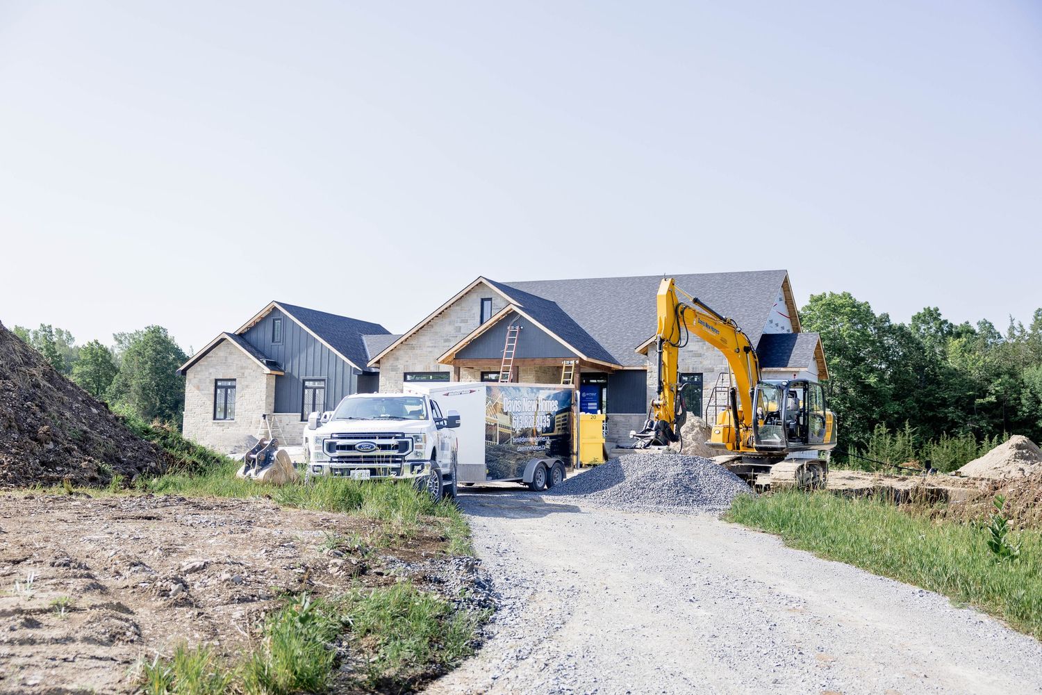 New home construction site with excavator, truck, and partially built house on a gravel driveway.