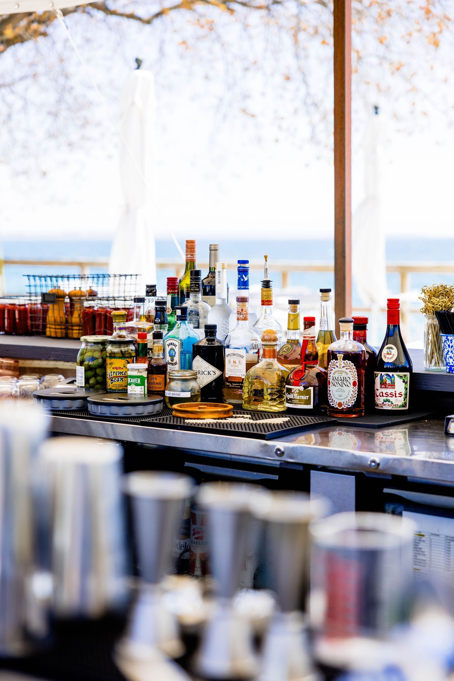 Fully stocked outdoor bar counter with liquor bottles and mixers with waterfront view in background.