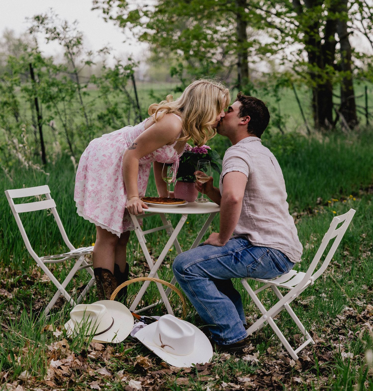 Couple kissing over a white outdoor bistro table with cowboy hats on the ground nearby.
