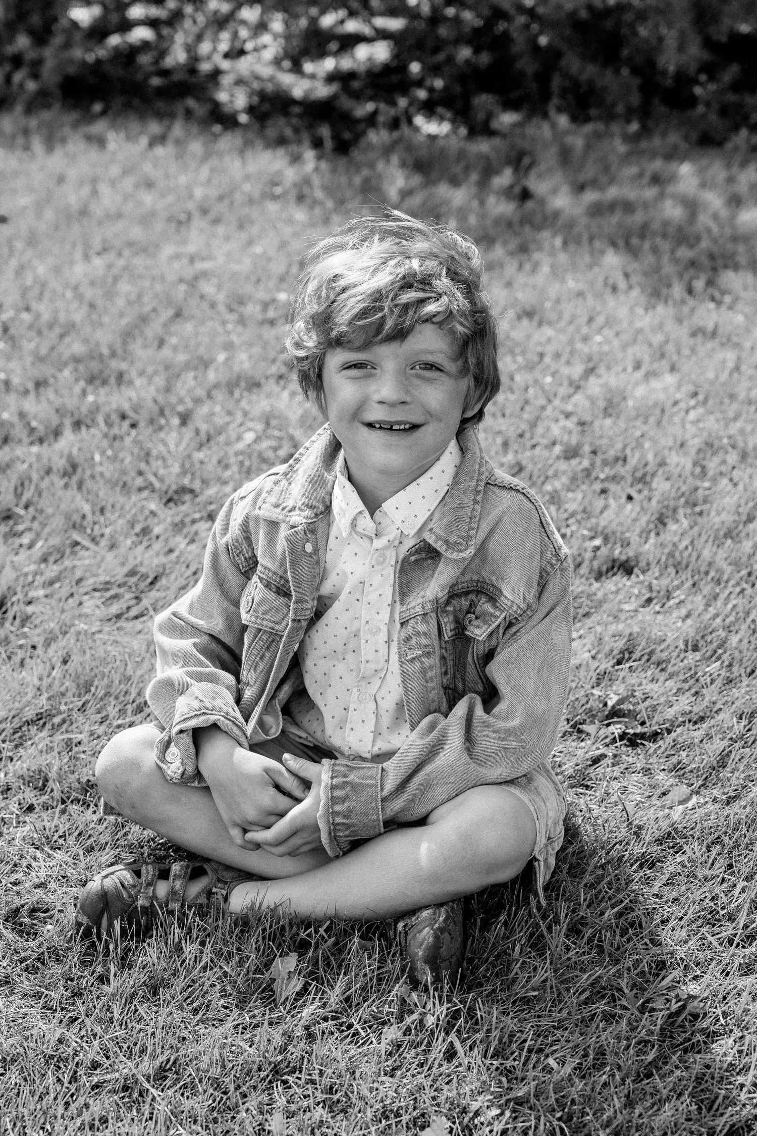 Black and white photo of a smiling young boy sitting cross-legged on grass wearing a denim jacket and shorts.