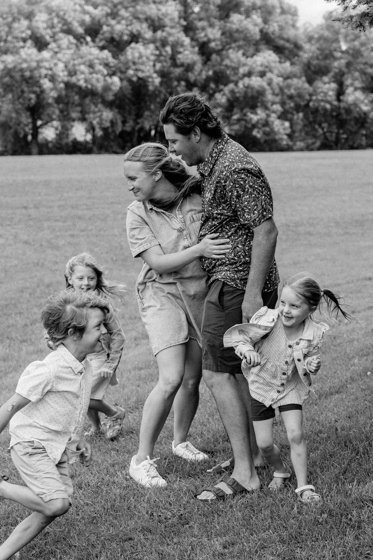 Black and white photo of a joyful family playing outdoors in a park, with children running toward smiling parents.