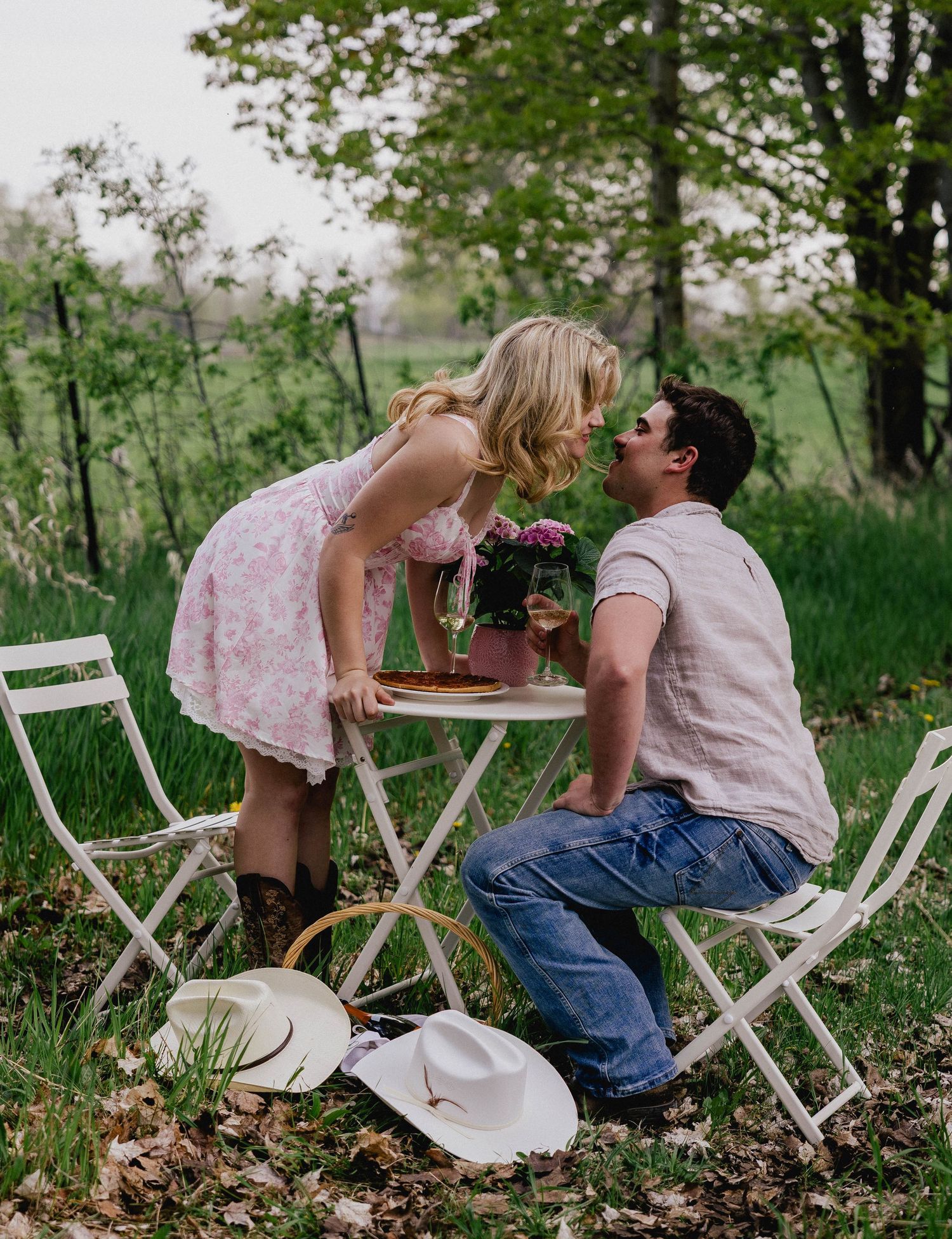 Couple sharing a romantic kiss over a white bistro table with flowers in a scenic outdoor setting.