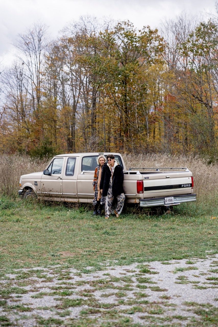 Two stylishly dressed women posing beside a vintage Ford pickup truck among autumn foliage.