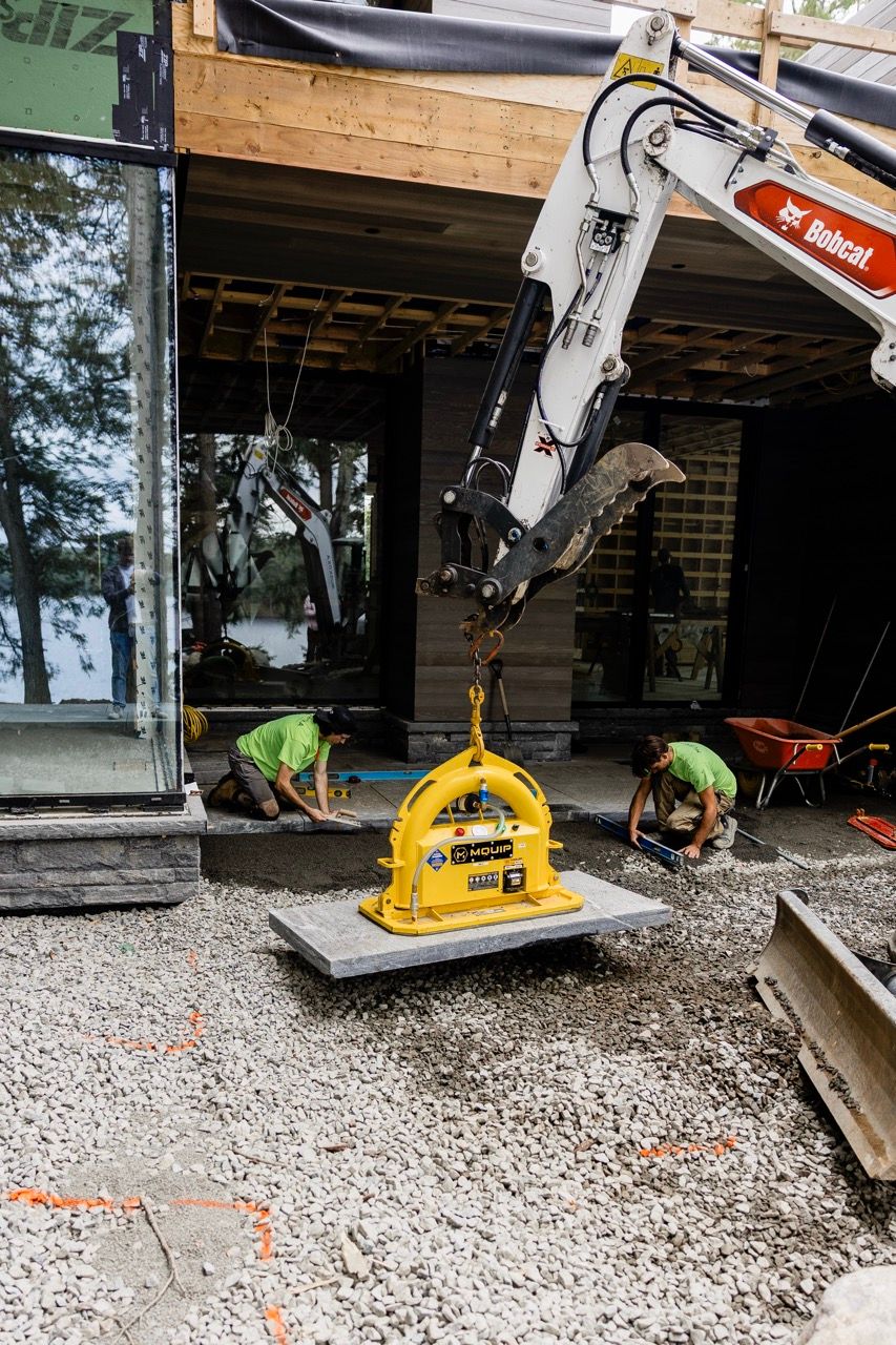 Bobcat excavator using a yellow MQUIP vacuum lifter to place stone slabs at a residential construction site.