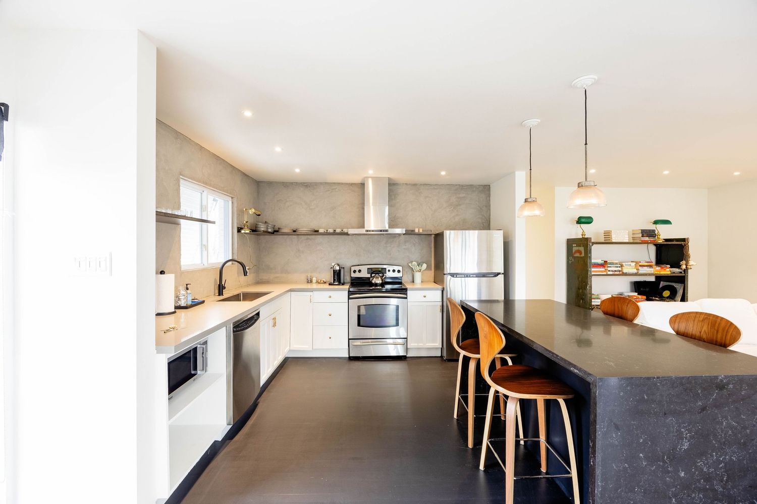 Modern white kitchen with dark island, bar stools, stainless steel appliances, and concrete backsplash.