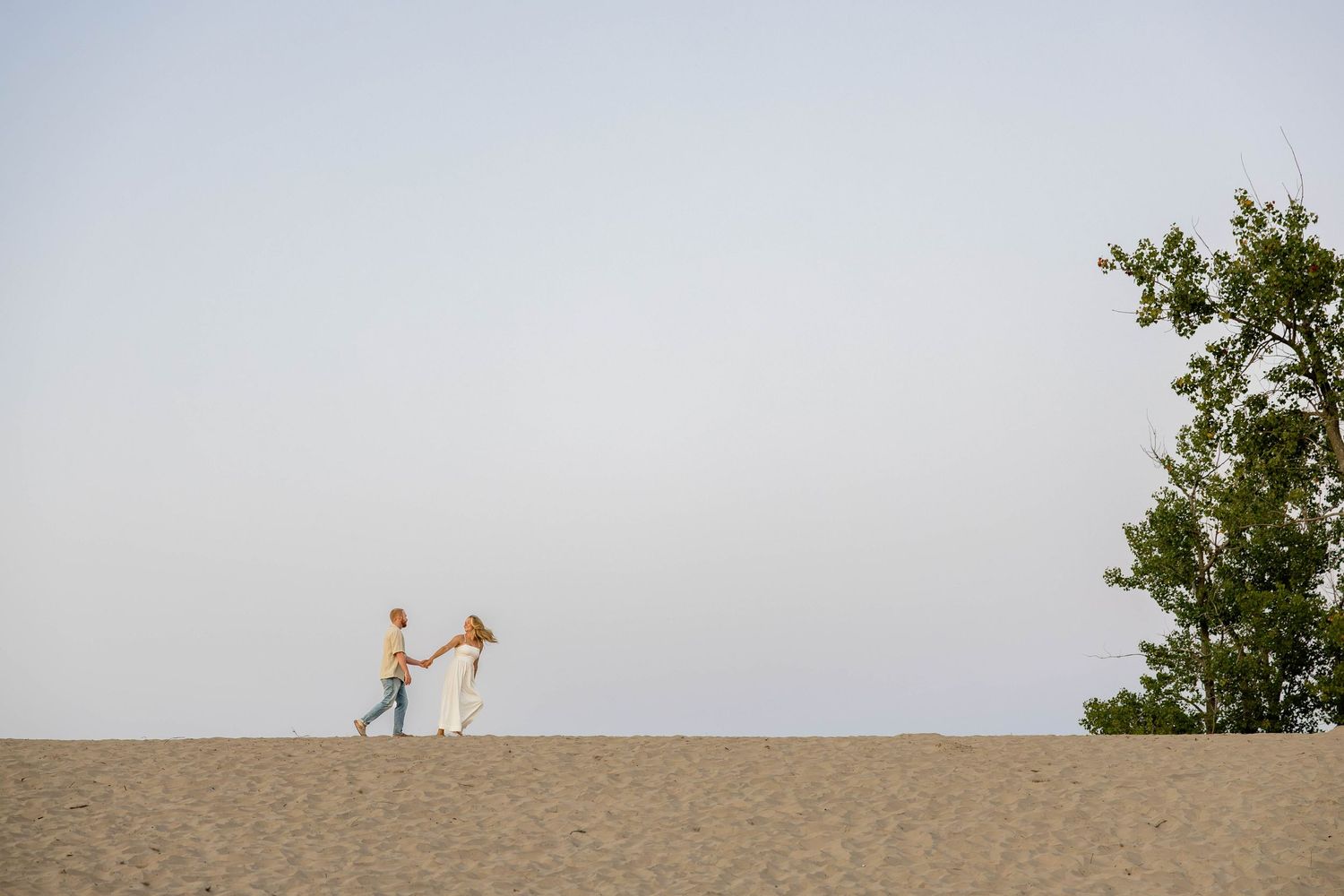 Couple dancing on sandy beach dunes with a tree and clear sky backdrop.