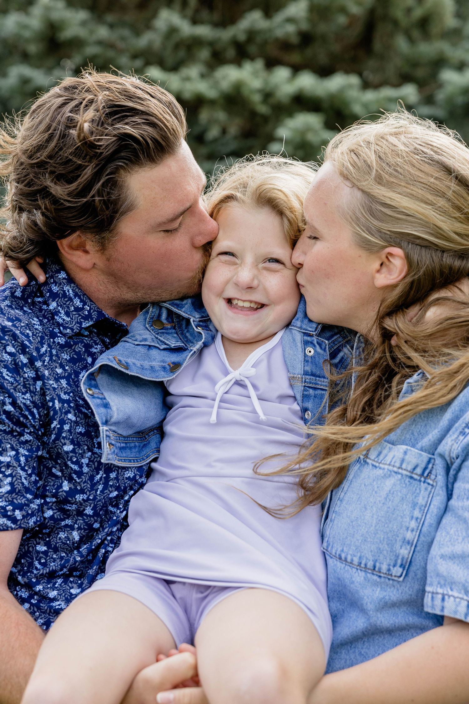 Parents kissing their smiling young daughter on both cheeks outdoors near green trees.