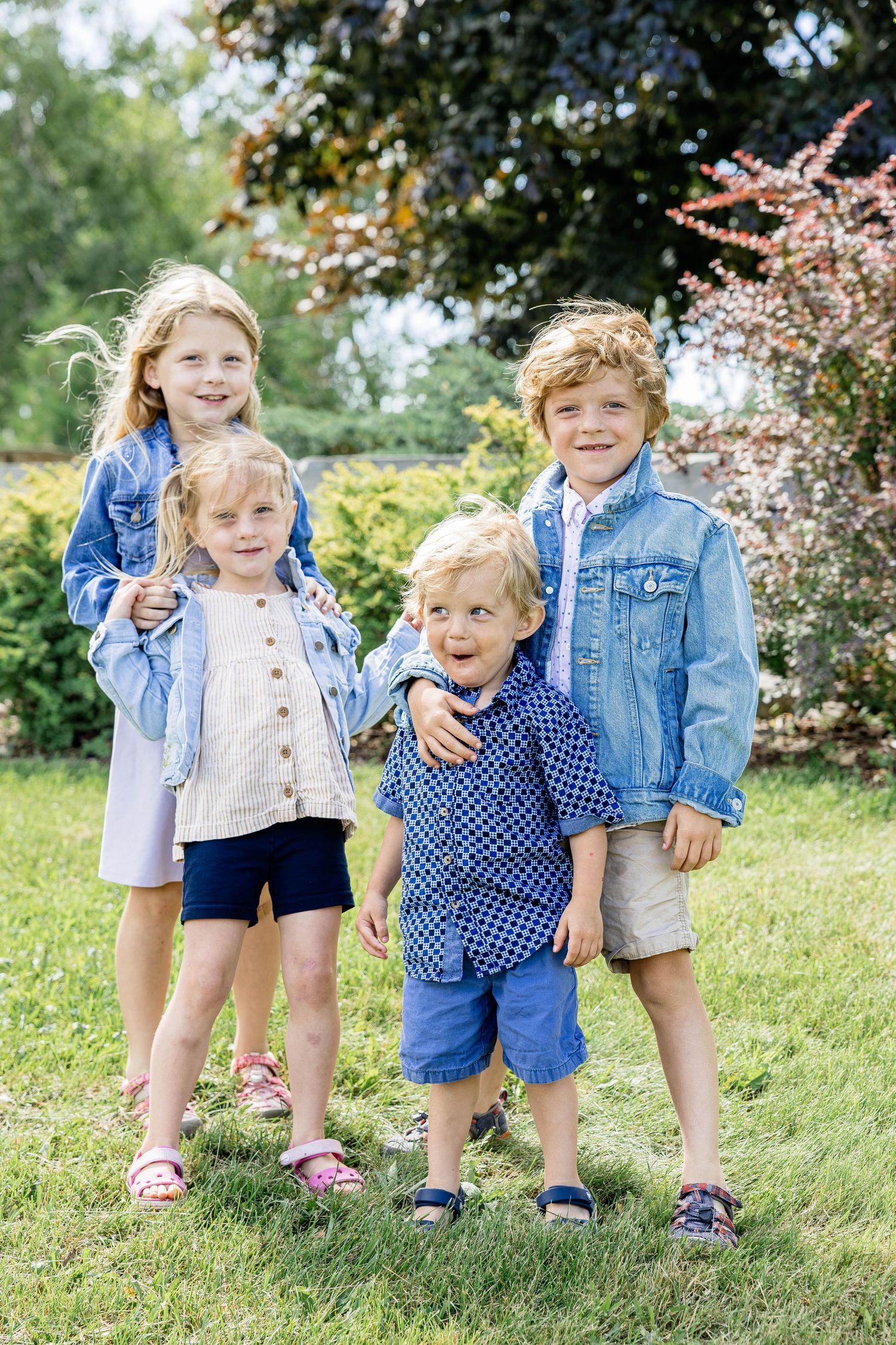 Four young children in denim jackets smile together outdoors on a sunny day in a lush green garden.