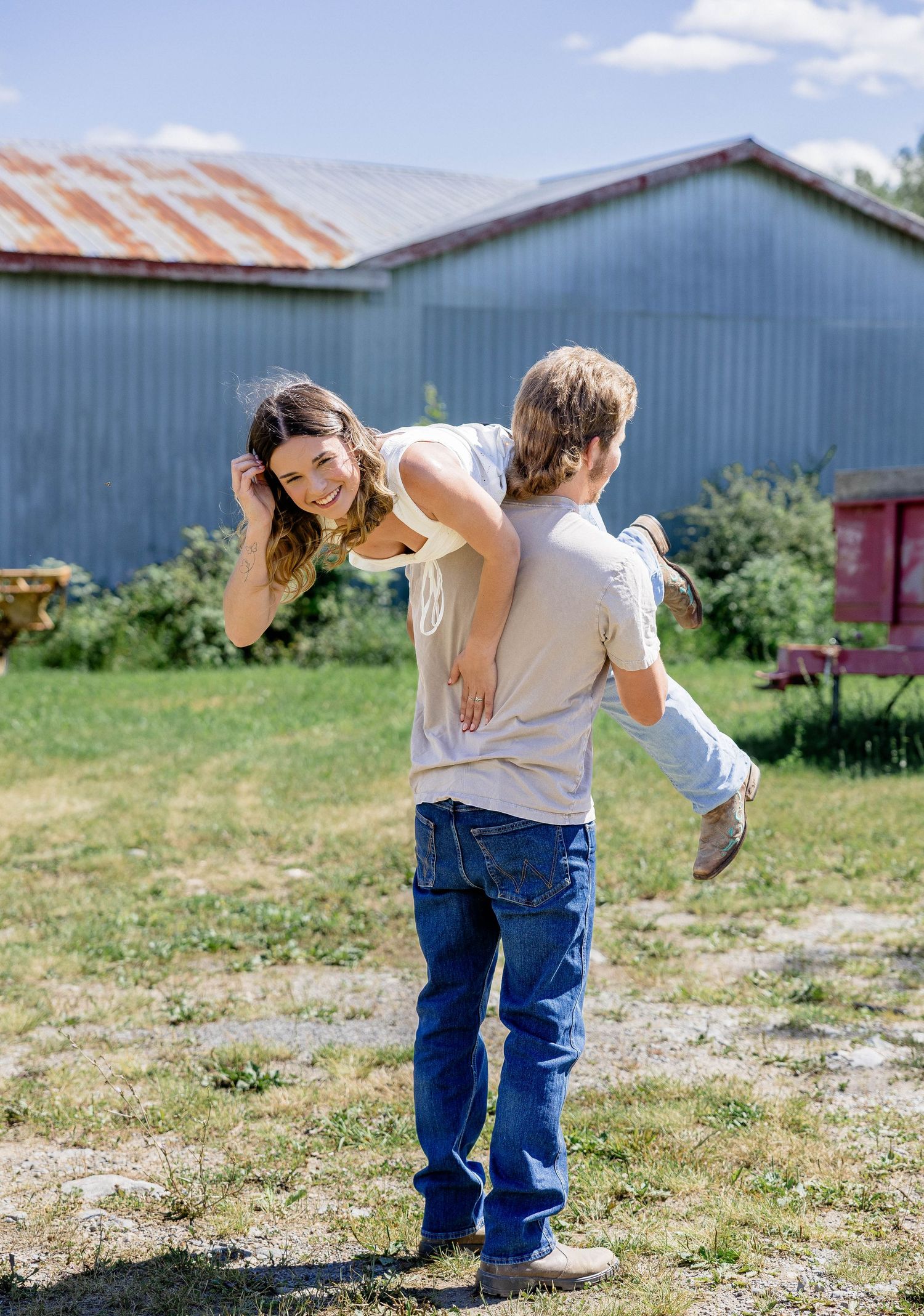 A young couple playing outdoors near a barn, man giving woman a piggyback ride on a sunny day.