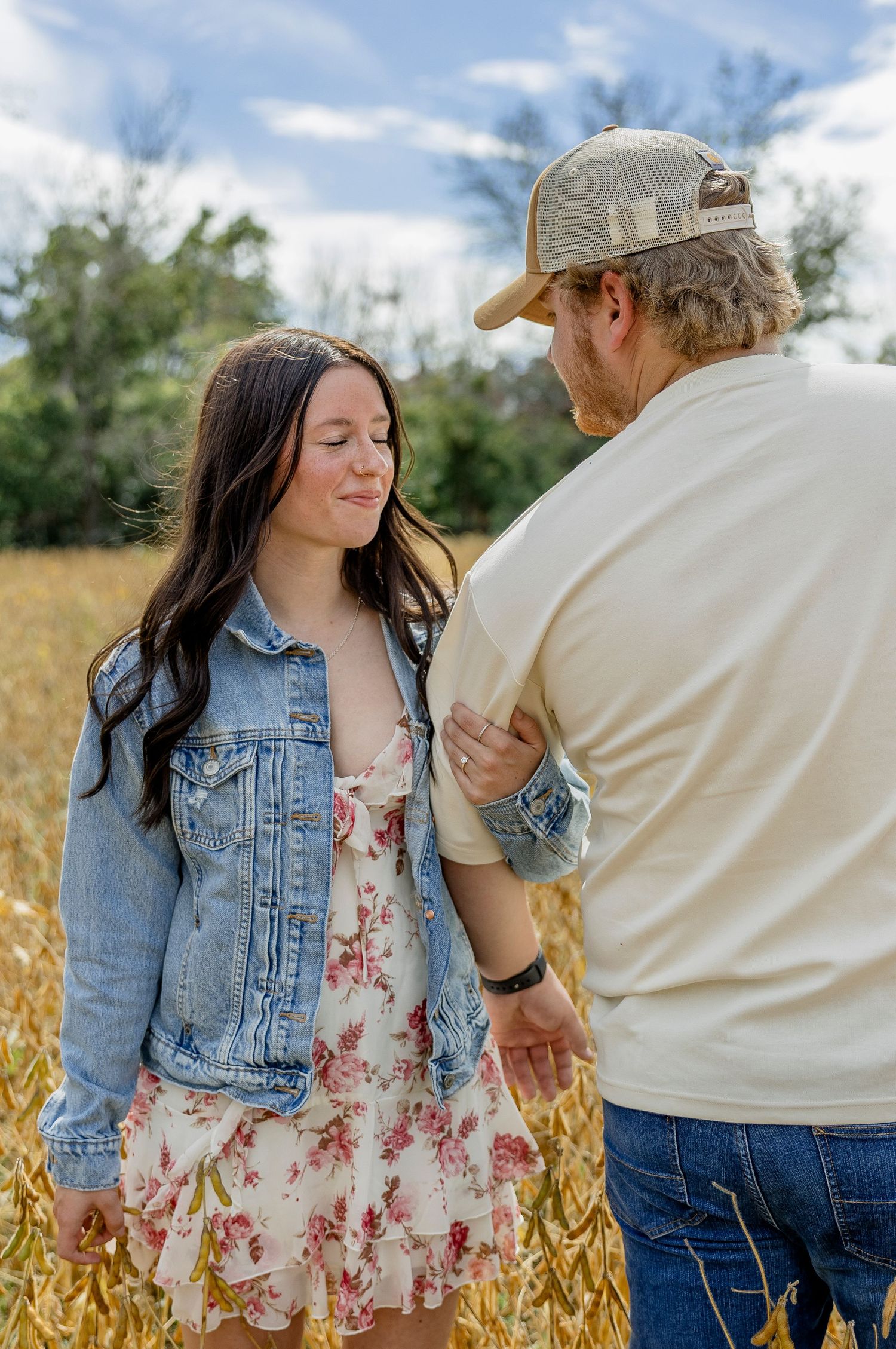 Couple standing in golden field, woman in floral dress and denim jacket, man in cap and cream polo shirt.