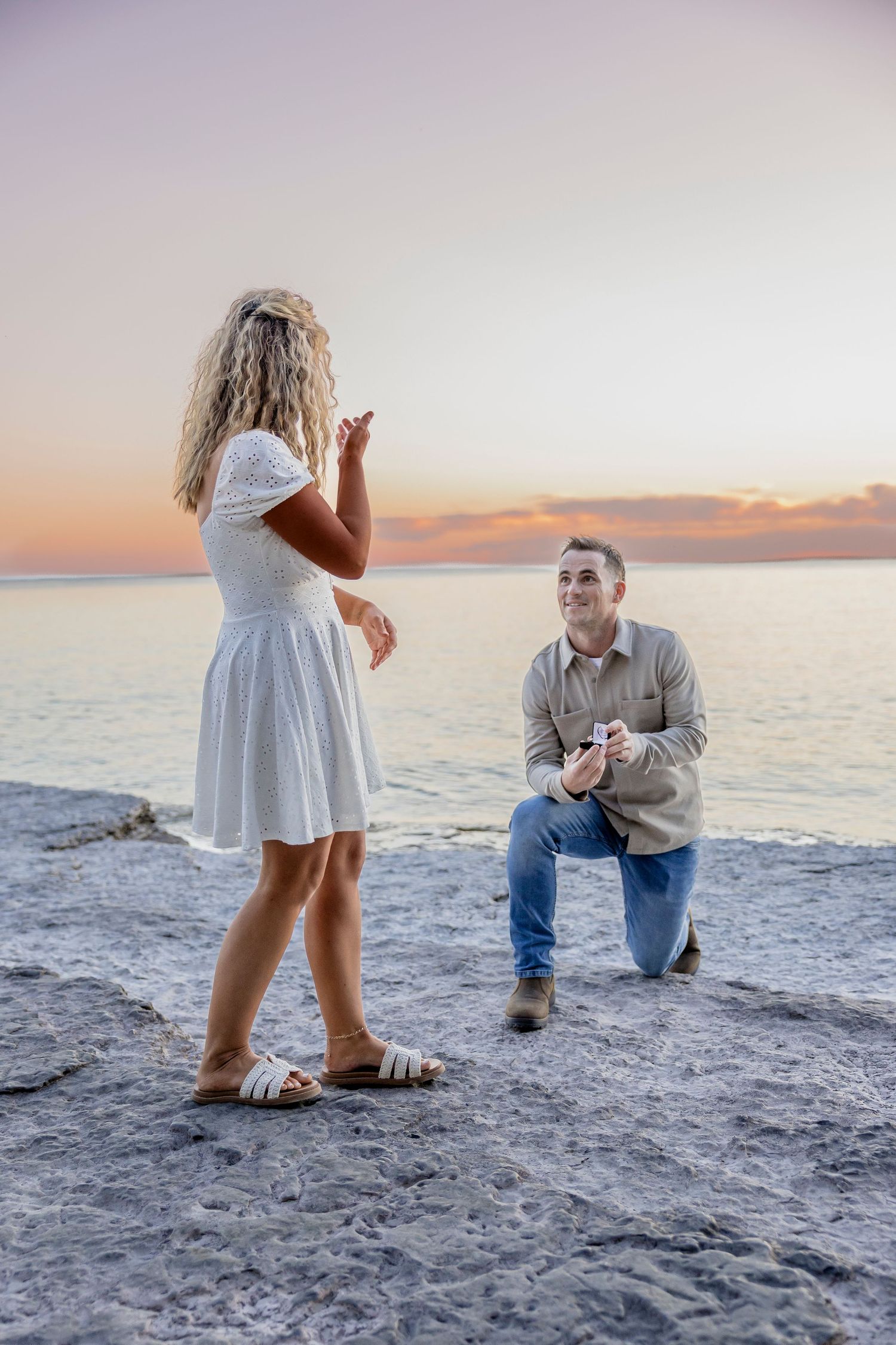A man proposes on one knee to a woman in a white dress at a rocky seaside sunset.