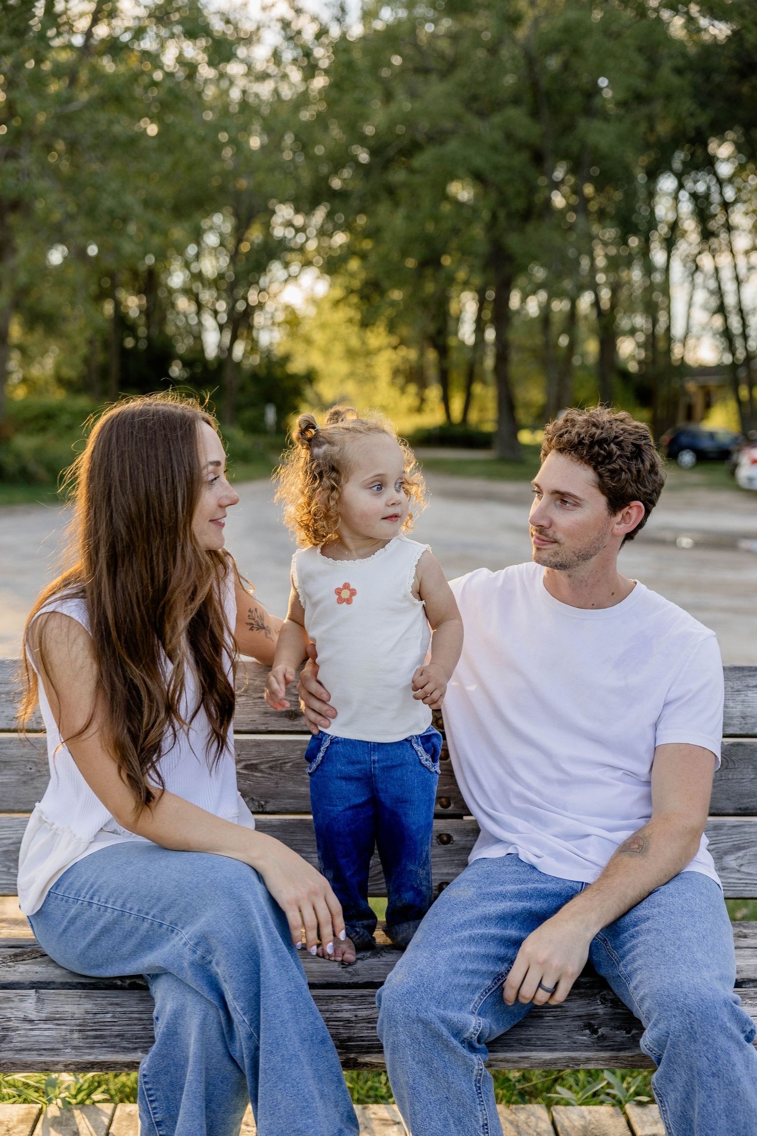 Young family with toddler sitting on a bench in a sunny park, parents in white tees and jeans smiling at child.