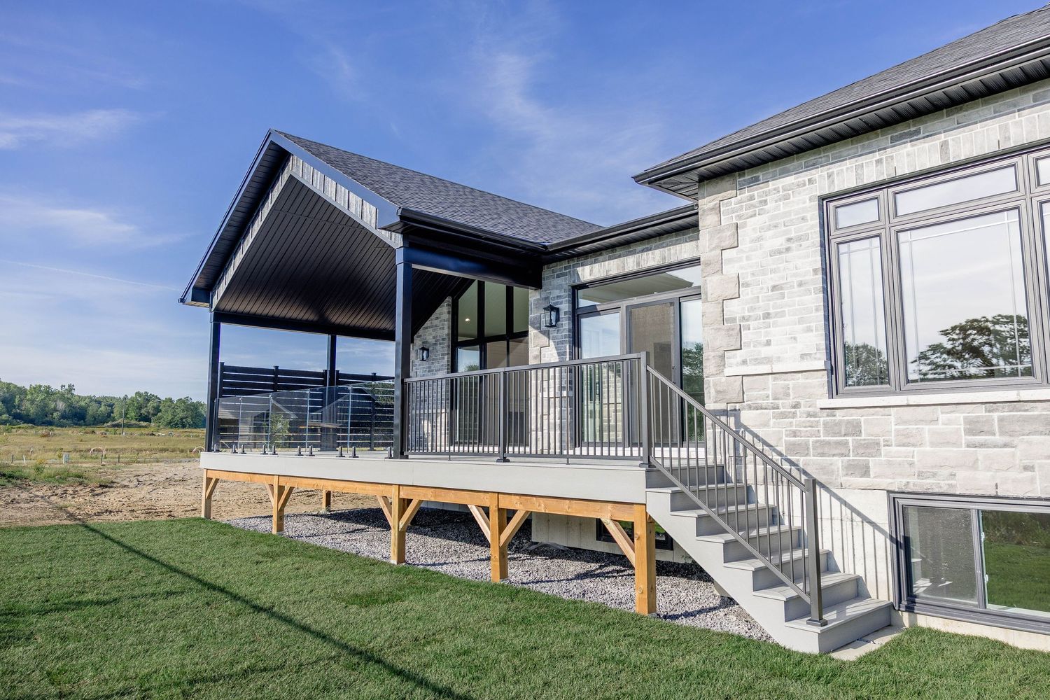 Modern stone home exterior featuring a covered deck with metal railings and wood stairs overlooking a green lawn.