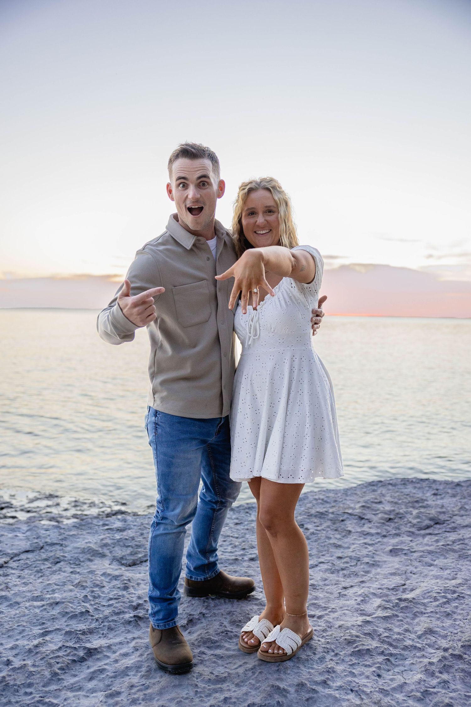 Excited couple at sunset beach showing engagement ring, man pointing while woman extends hand forward.