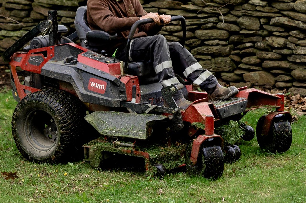 Person riding red Toro zero-turn lawn mower along stone wall on grassy landscape.