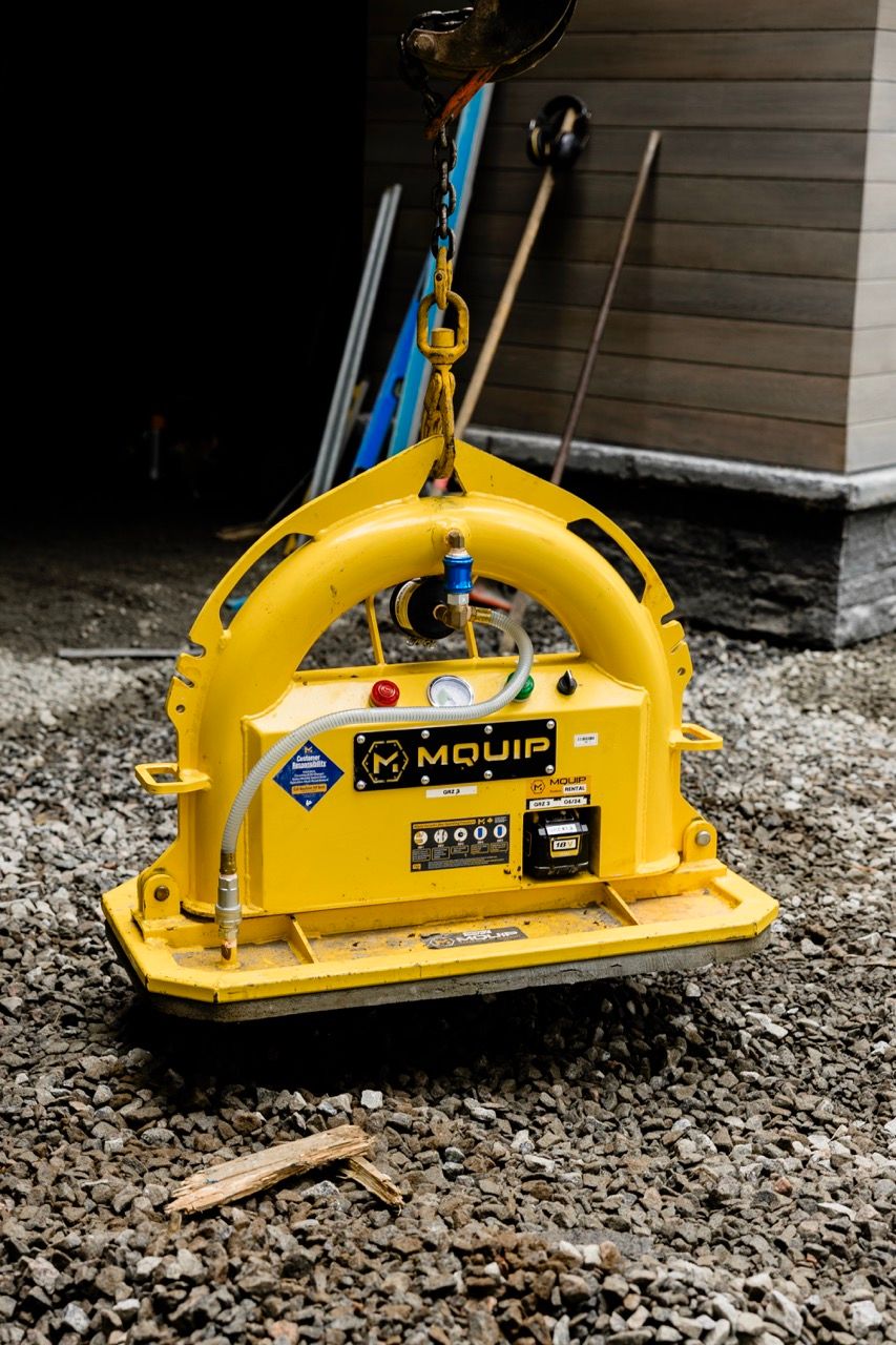 Yellow MQUIP vacuum lifting attachment suspended by crane hook on gravel at construction site.