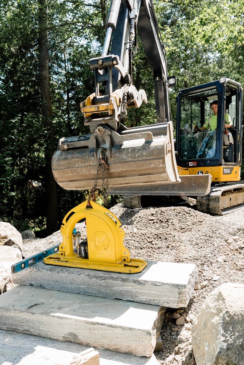 CAT mini excavator with yellow vacuum stone lifter lifting granite slab at construction site.