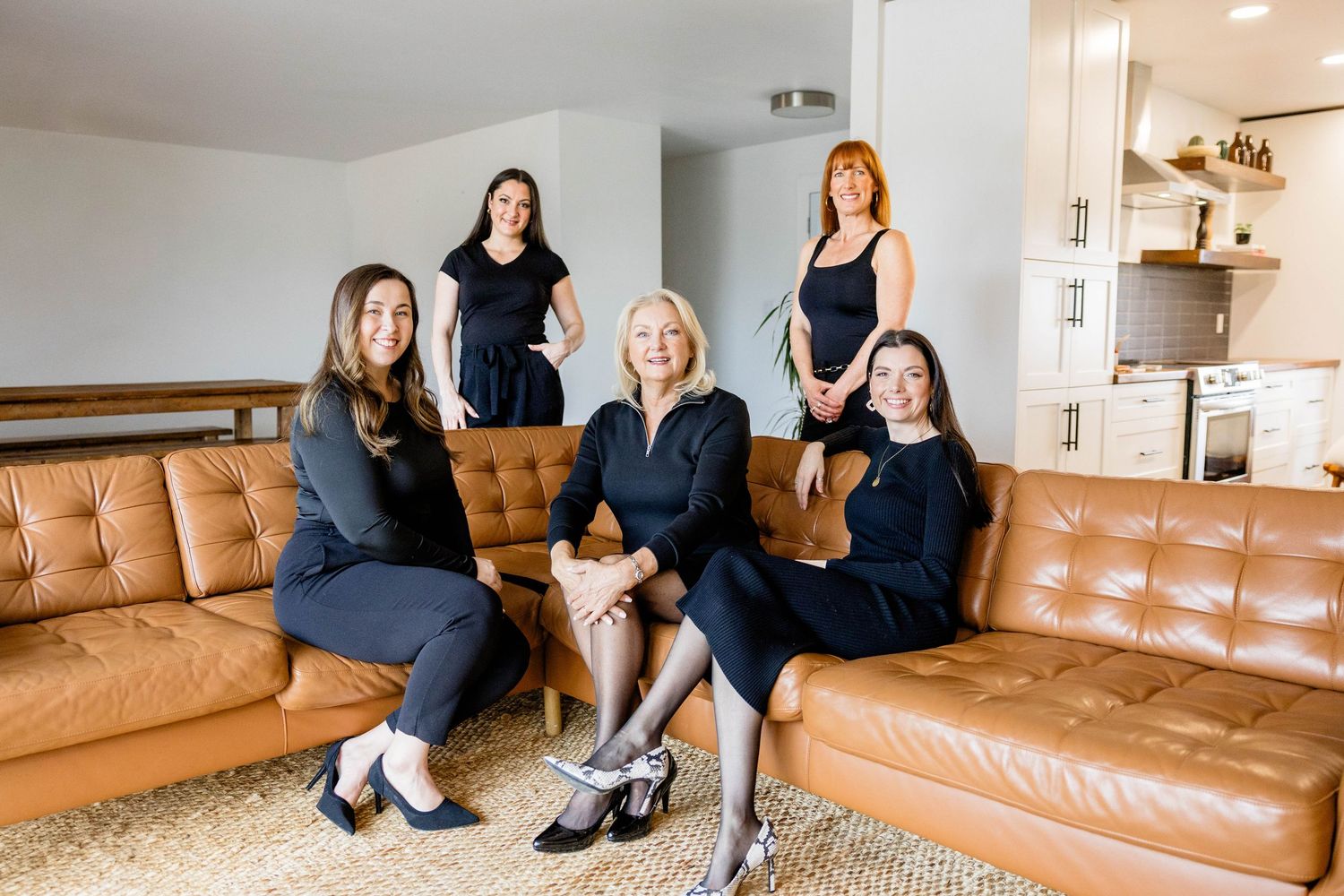 Five professional women in black attire posing together in a stylish modern living room.