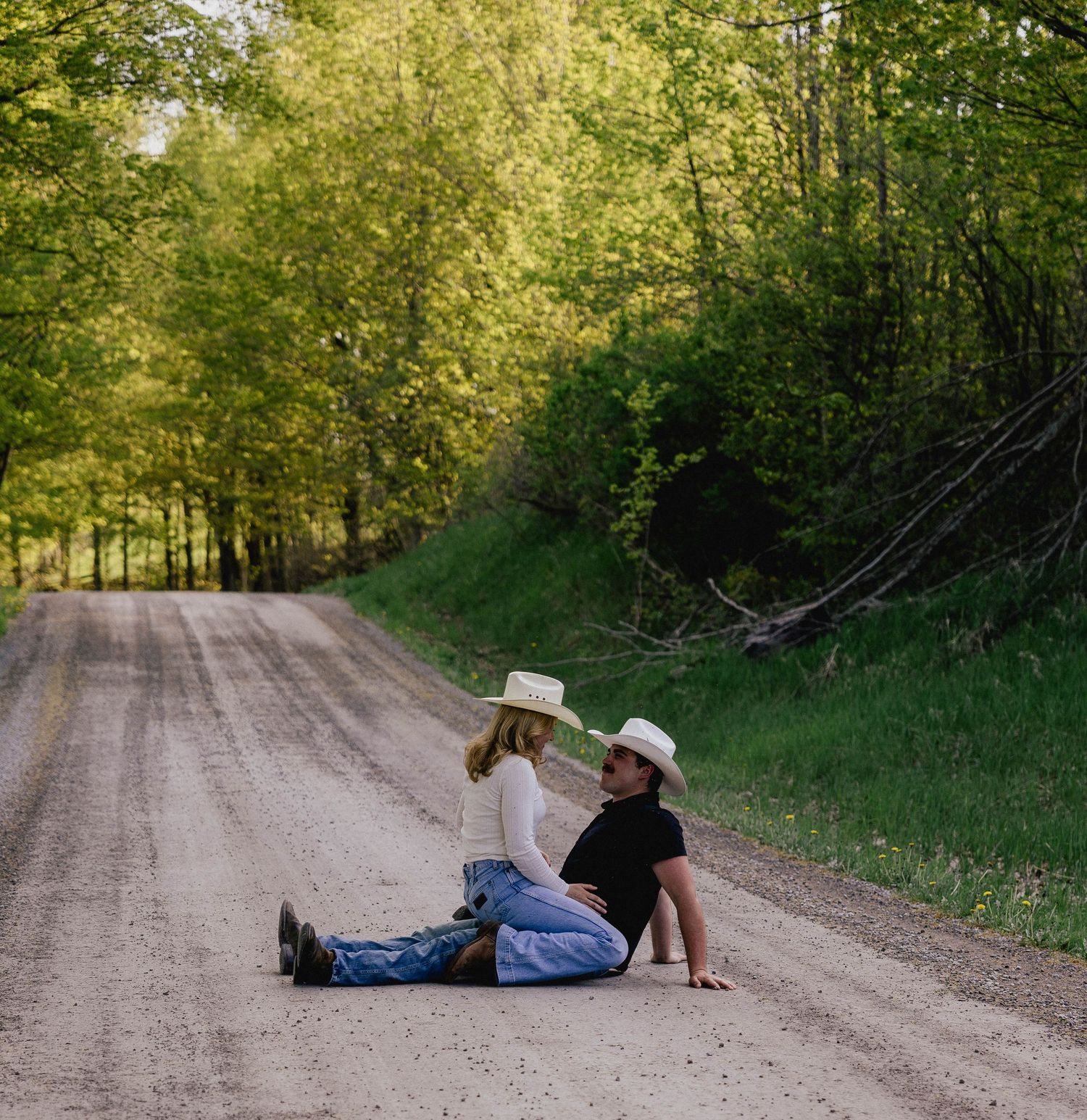 Couple sitting close on rural dirt road, wearing cowboy hats, surrounded by green tree-lined path.