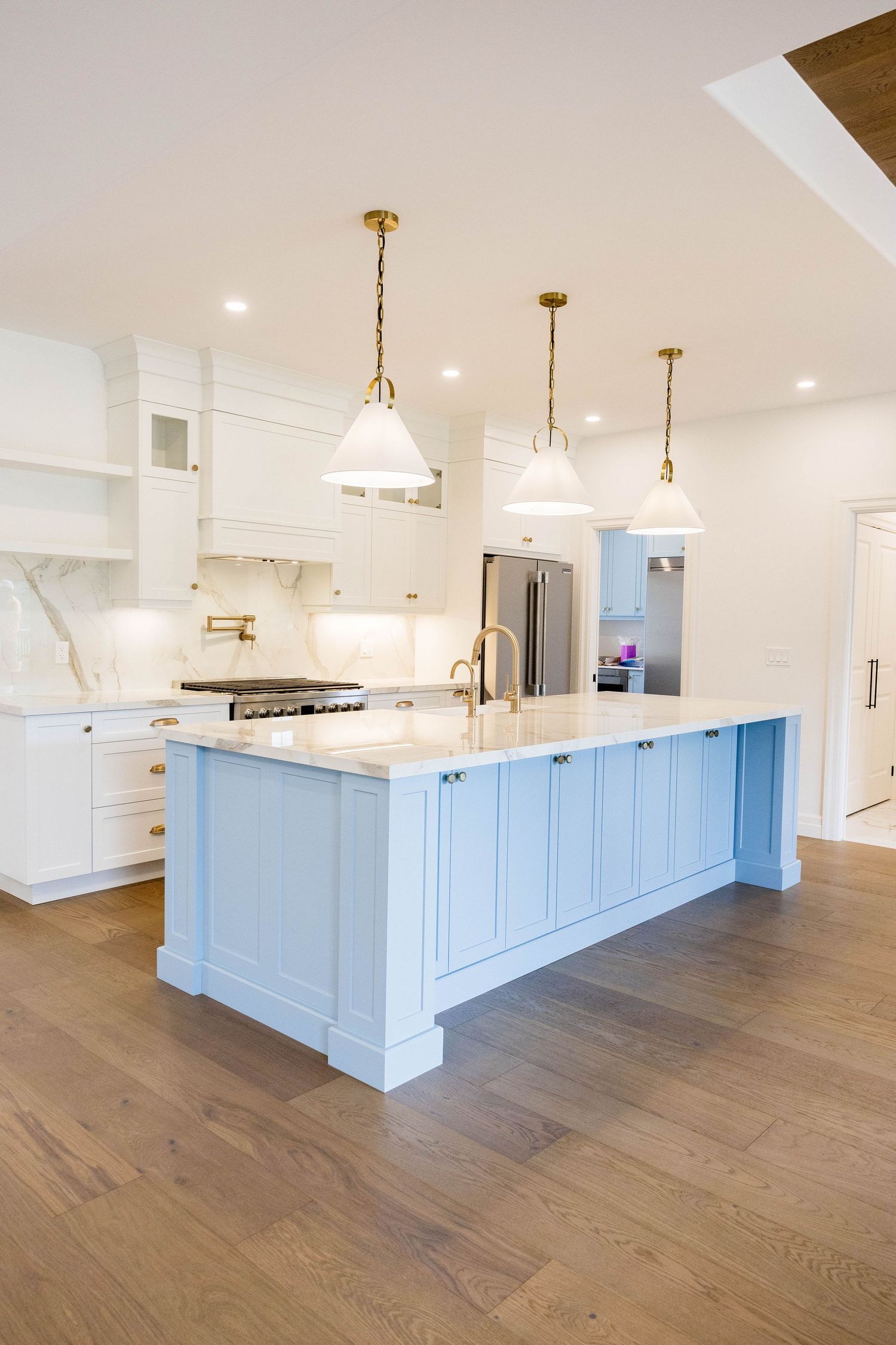 Bright white kitchen with light blue island, gold pendant lights, marble countertops, and hardwood floors.
