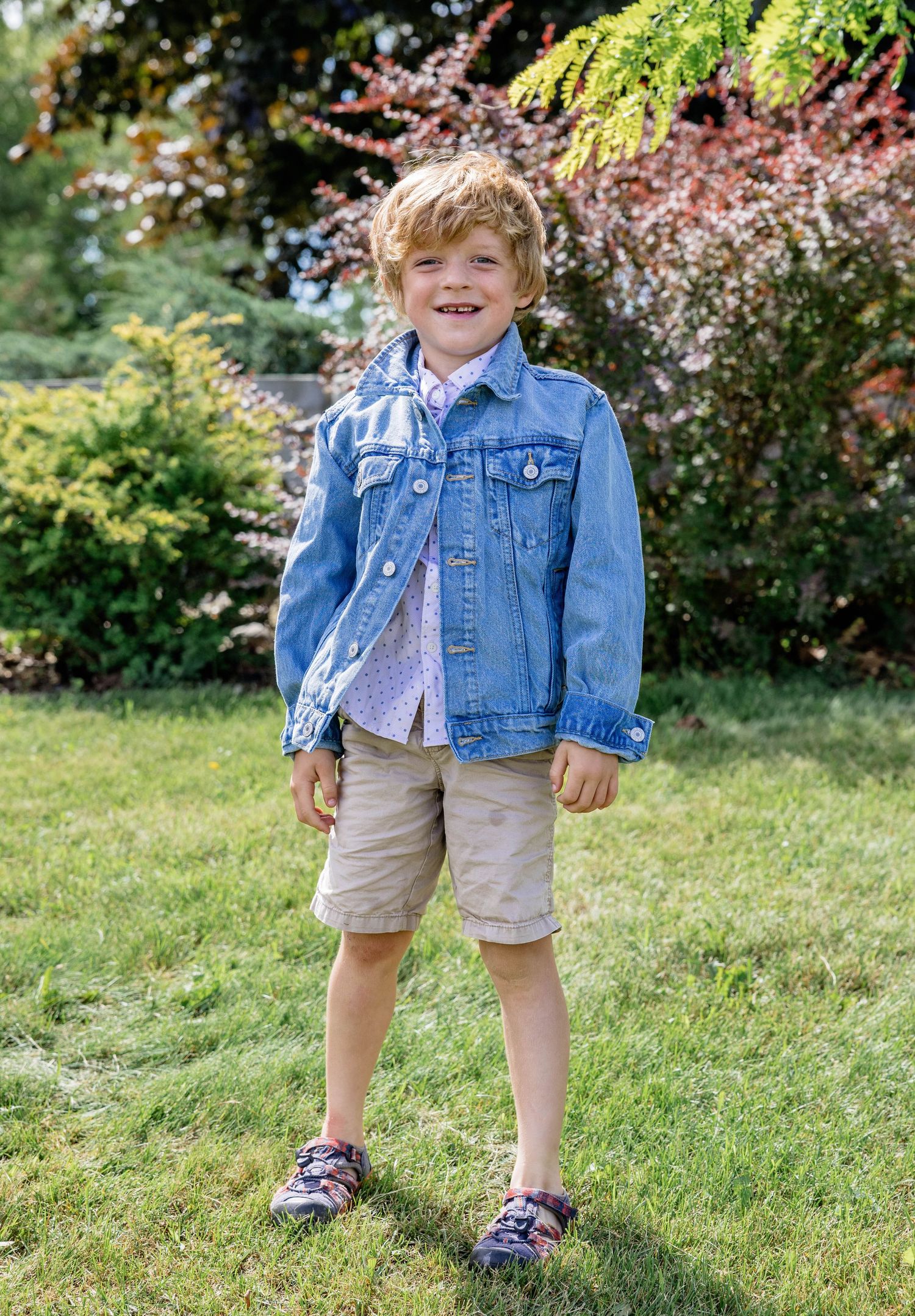 Smiling young boy in denim jacket, floral shirt, khaki shorts, and sandals standing on green grass outdoors.