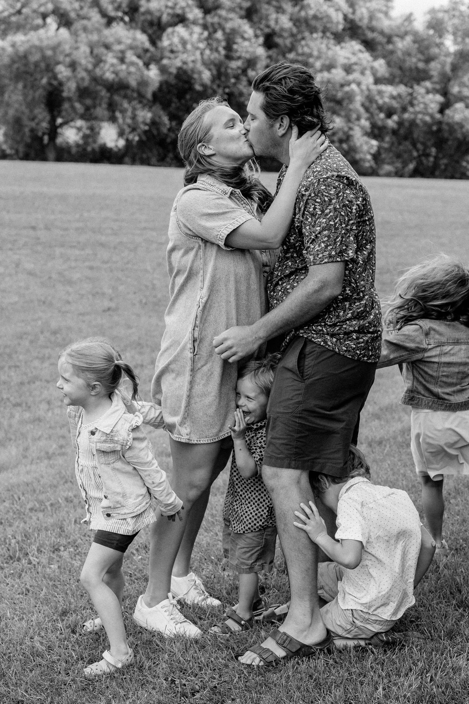 Black and white photo of a couple kissing in a park surrounded by their young children playing around them.