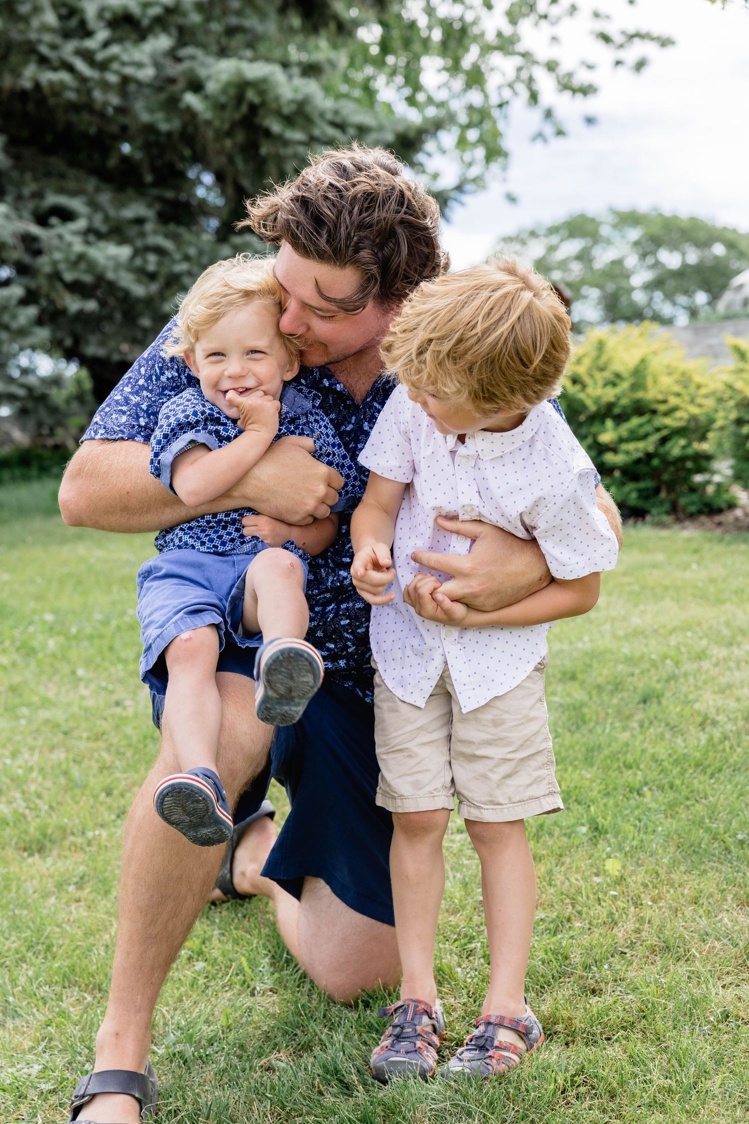 A father kneels on grass hugging two young blonde boys in a lush green backyard on a summer day.
